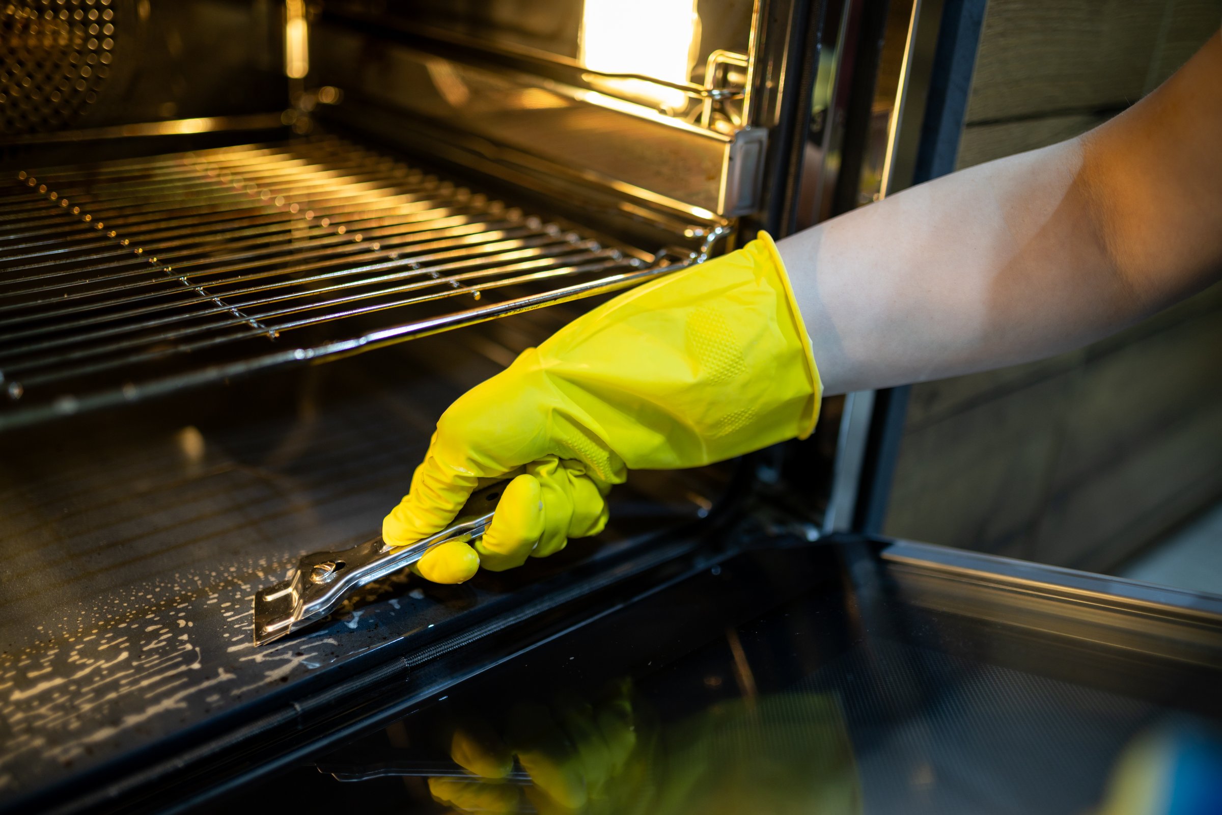 A hand wearing a yellow glove is using a scraper to clean the inside of an oven. The focus is on removing grime and grease from the oven's surface.