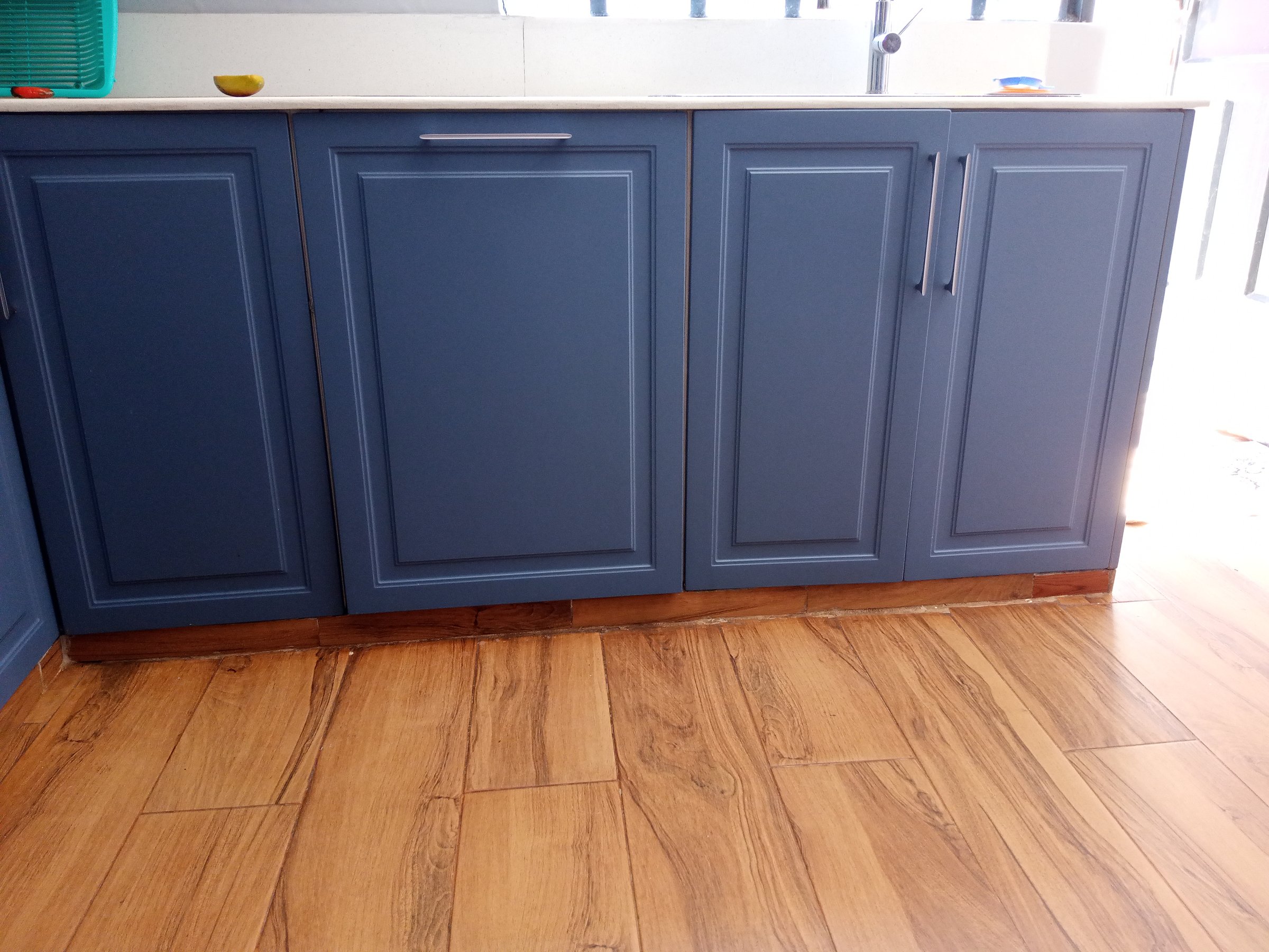 Blue kitchen cabinets with decorative panels and wooden flooring, partially illuminated by natural light.