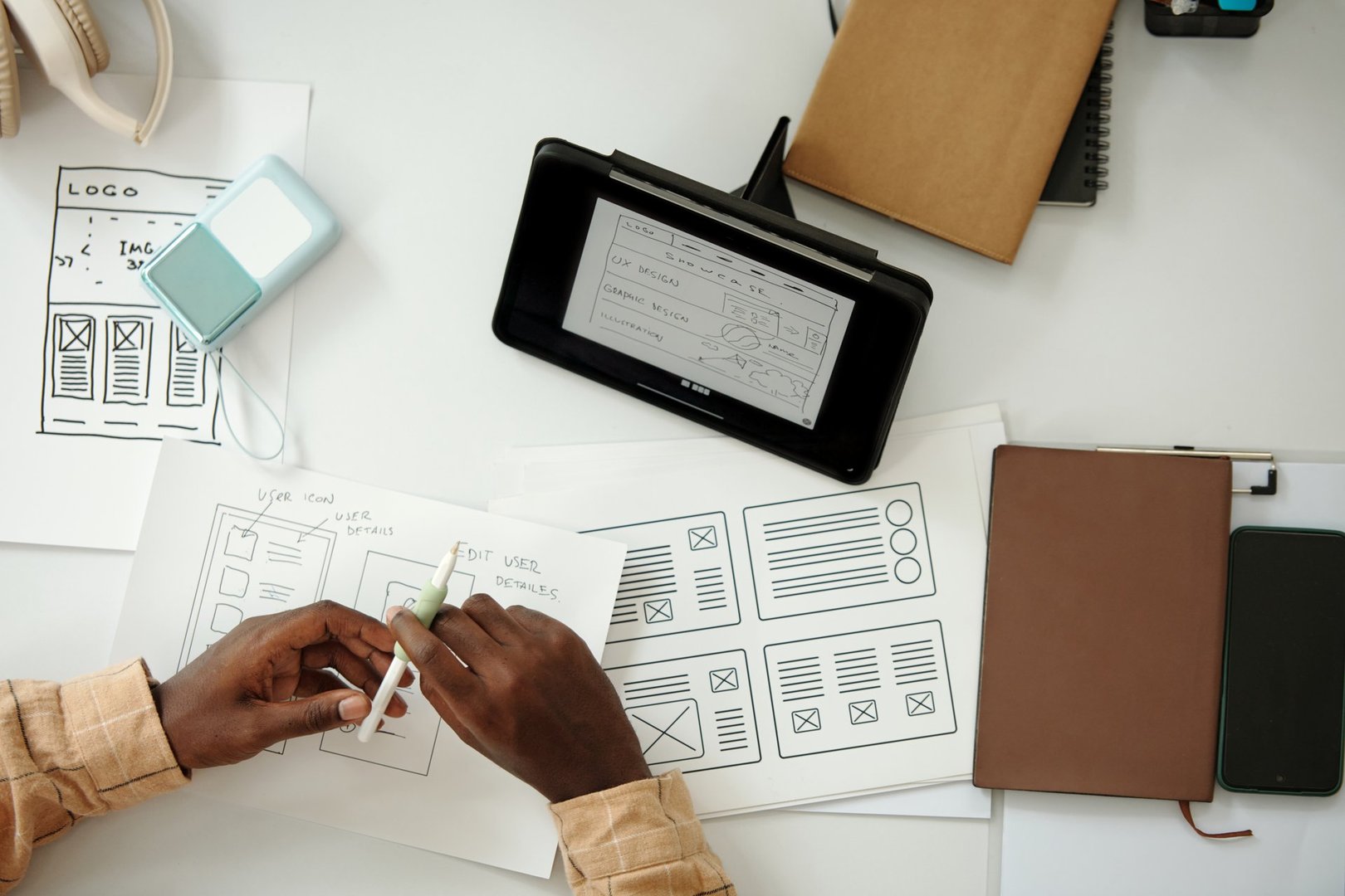 Hands sketching wireframes on paper with tablet displaying digital prototype. Workspace includes notebooks, pens, and digital gadgets for design process