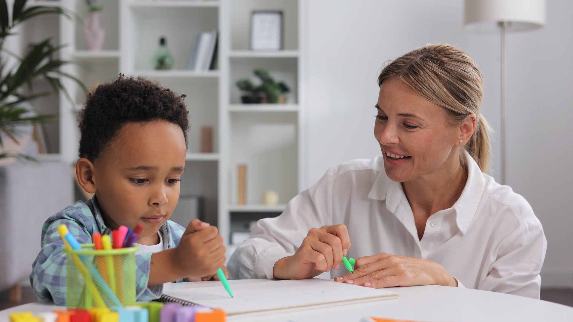 Adorable little african american boy with curly hair having fun at pediatric specialist appointment, happy child drawing with female therapist, exercises for children with autism.