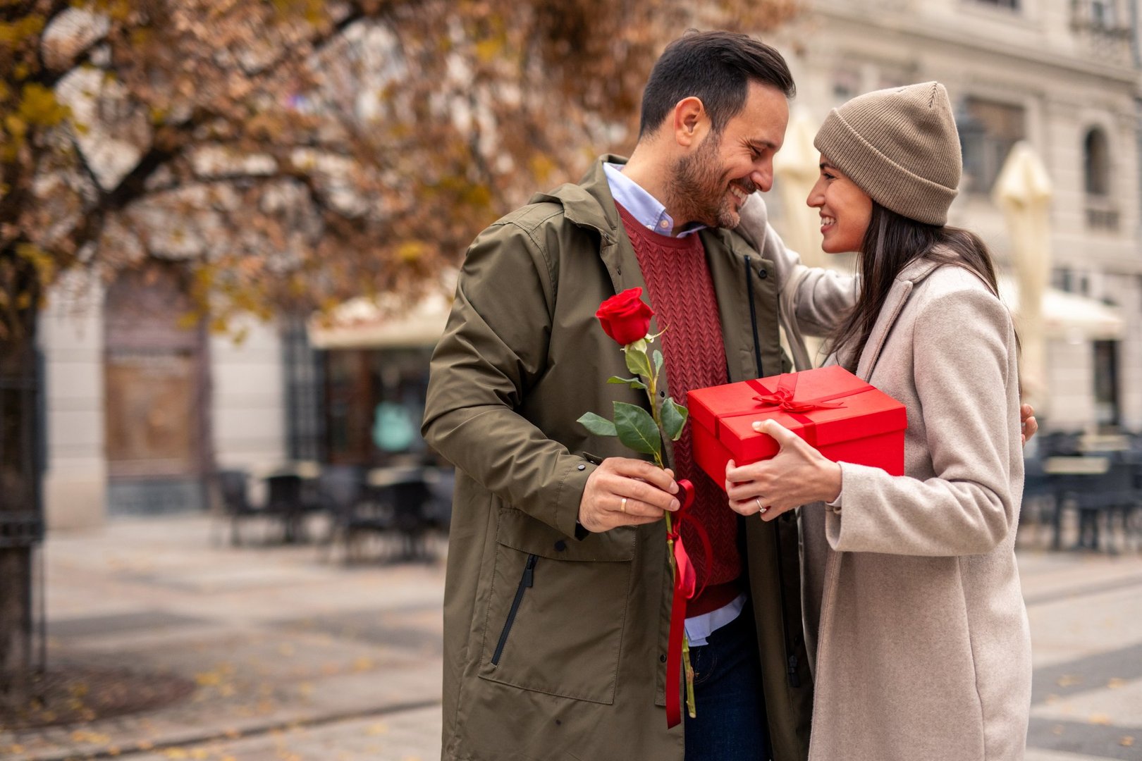 Mid adult Caucasian male surprises female partner with a wired rose on a bustling city street. Both dressed in cozy coats, sharing smiles, creating joyful moments. A red gift box adds charm to this lively urban setting.