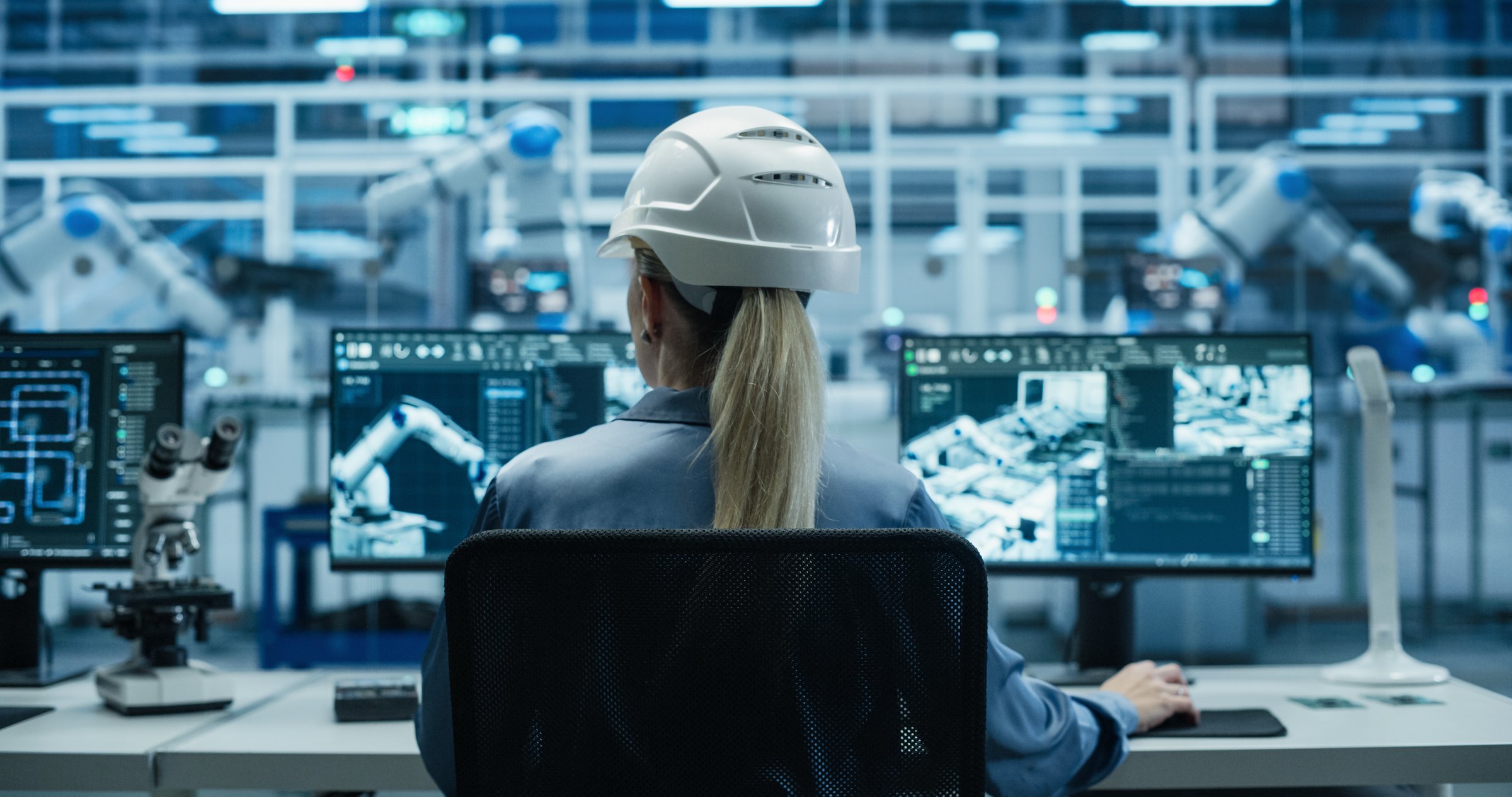 Female Industrial Engineer Working Behind a Desk in a Modern Manufacturing Facility with Automated Robotic Arms. Specialist Monitoring Circuit Board Assembly Line and Optimizing Production Output