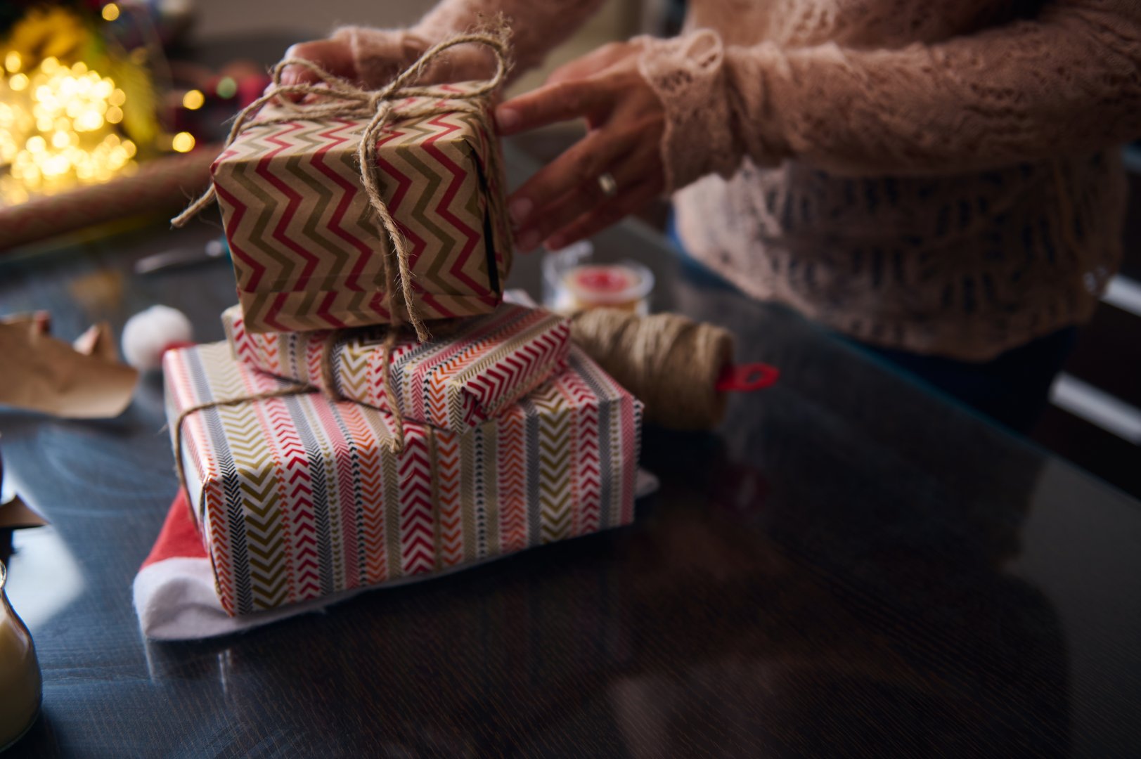 A close-up of hands wrapping beautifully patterned gifts, highlighting holiday preparation and care. Surrounded by festive decorations, twine, and wrapping materials, this scene embodies the joy of giving.