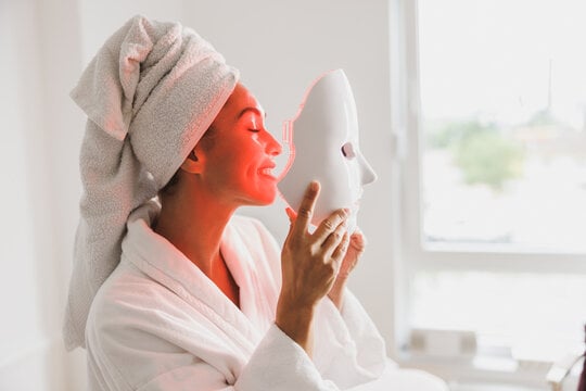 Person in a bathrobe with a towel on their head holds a light therapy mask, smiling, near a bright window.