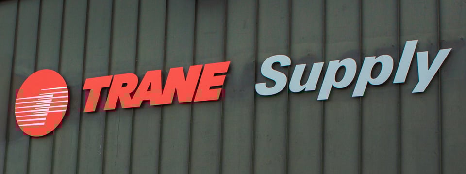 Trane Supply logo on a building wall, featuring a red and white circular emblem next to the brand name in red and gray letters.