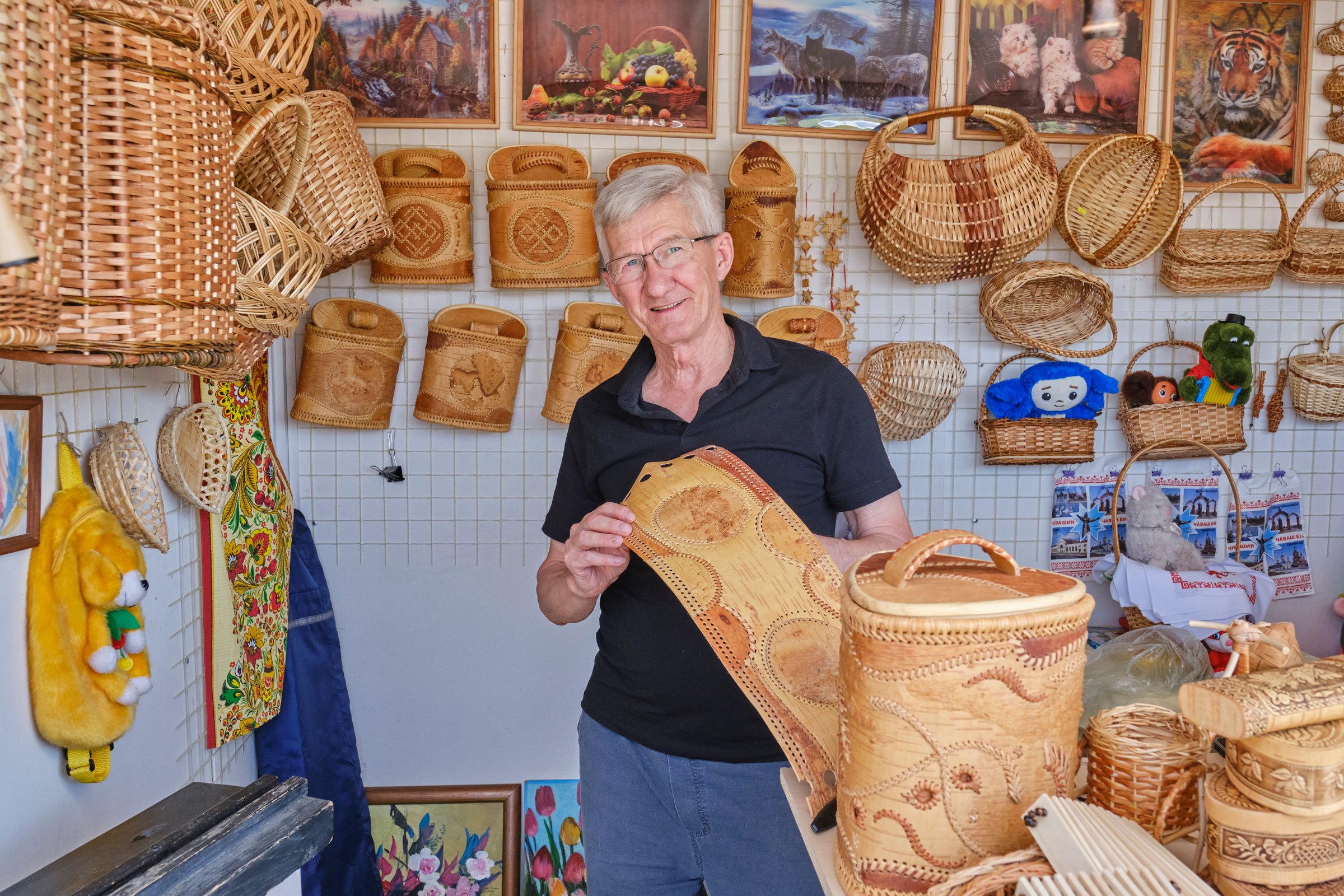 Cheboksary, Russia - June 11, 2024: Senior artisan proudly presenting intricately handcrafted birch bark items and woven baskets in craft stall surrounded by traditional handmade products.