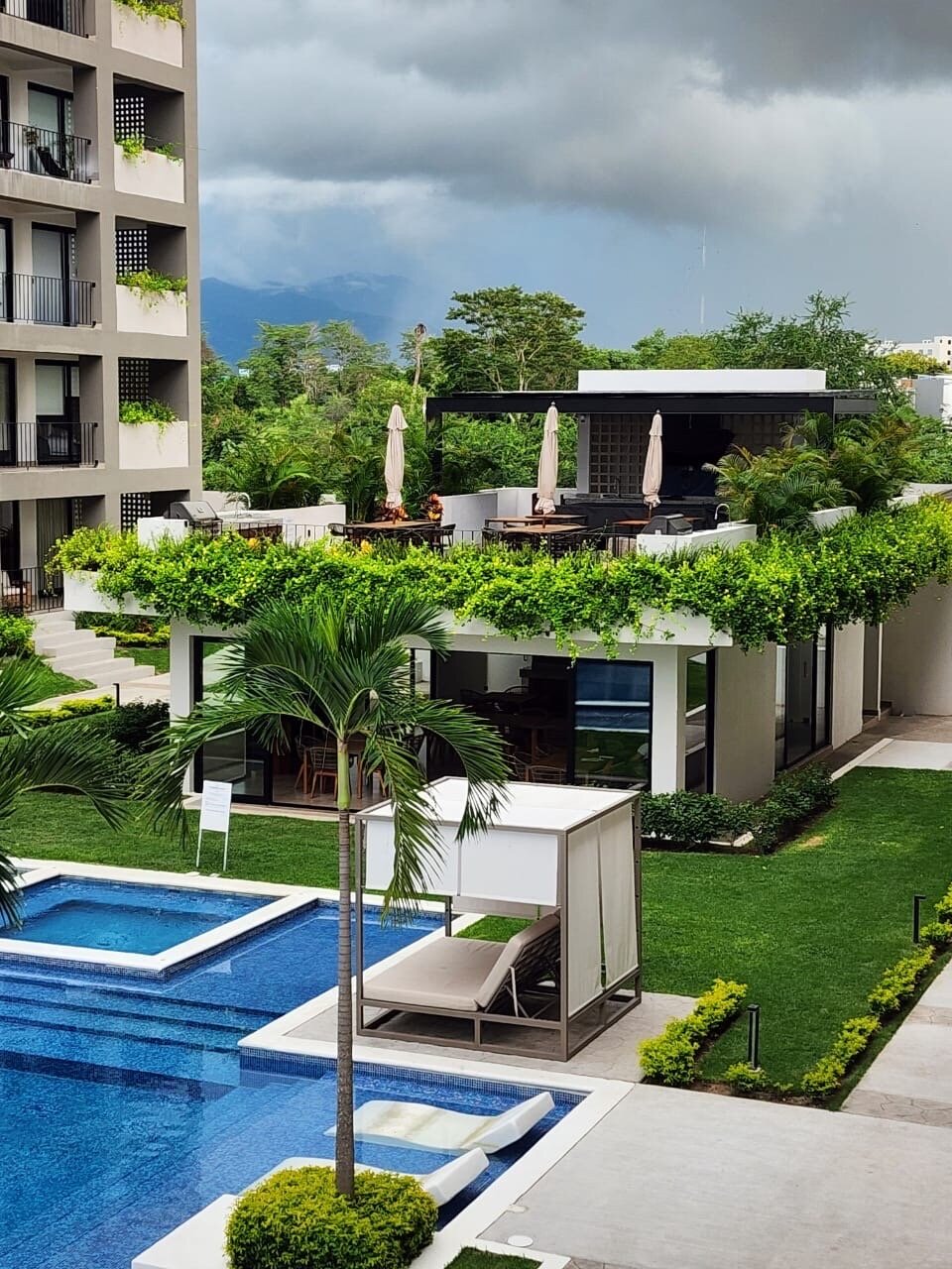 Modern apartment complex with a rooftop lounge, swimming pool, and landscaped garden, set against a cloudy sky.
