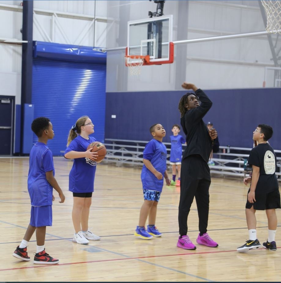 A group of kids watch a man shoot a basketball in an indoor court. They are all wearing casual sports attire.