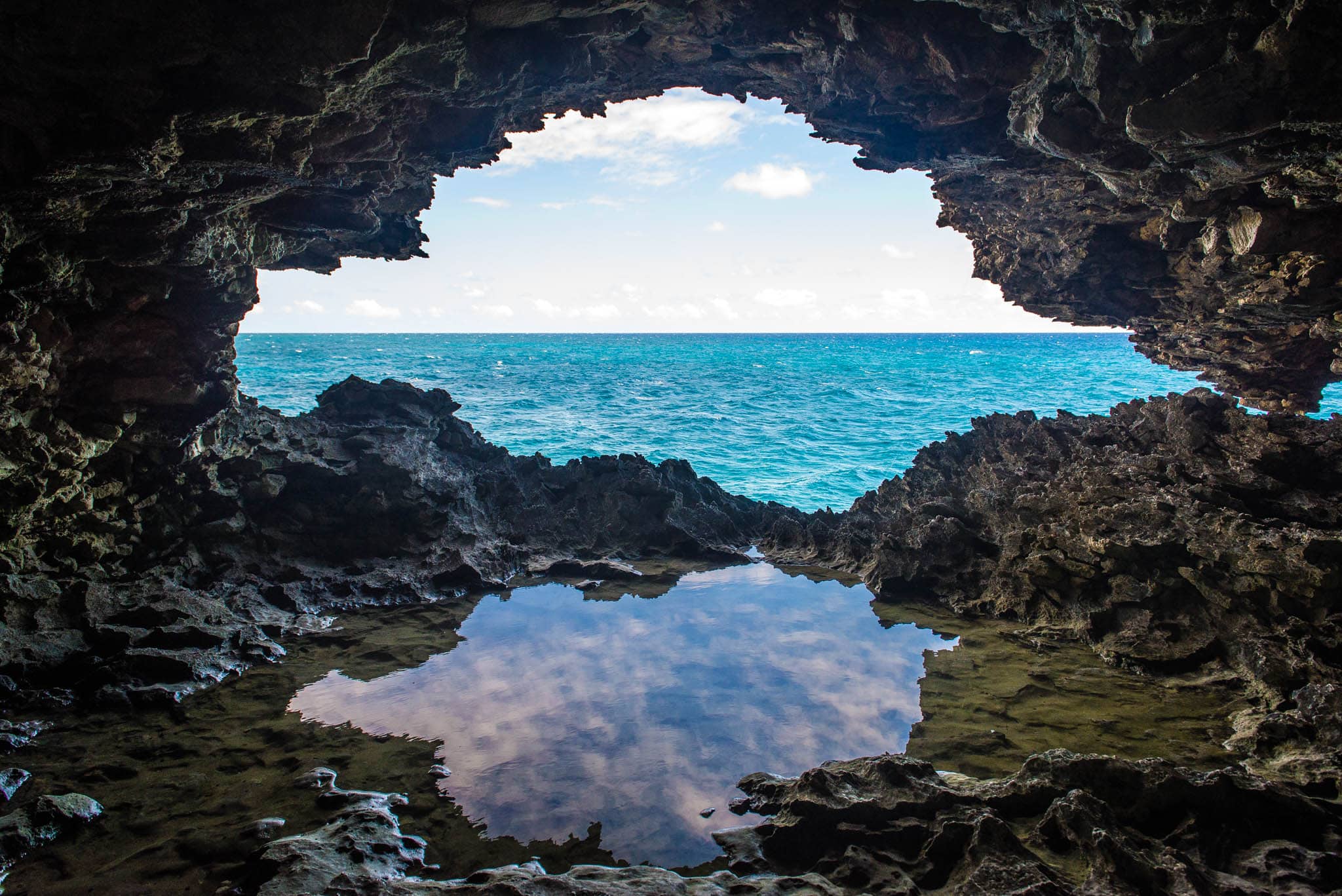 View from inside a rocky cave, looking out at the ocean through an opening with a reflection in a small water pool inside.