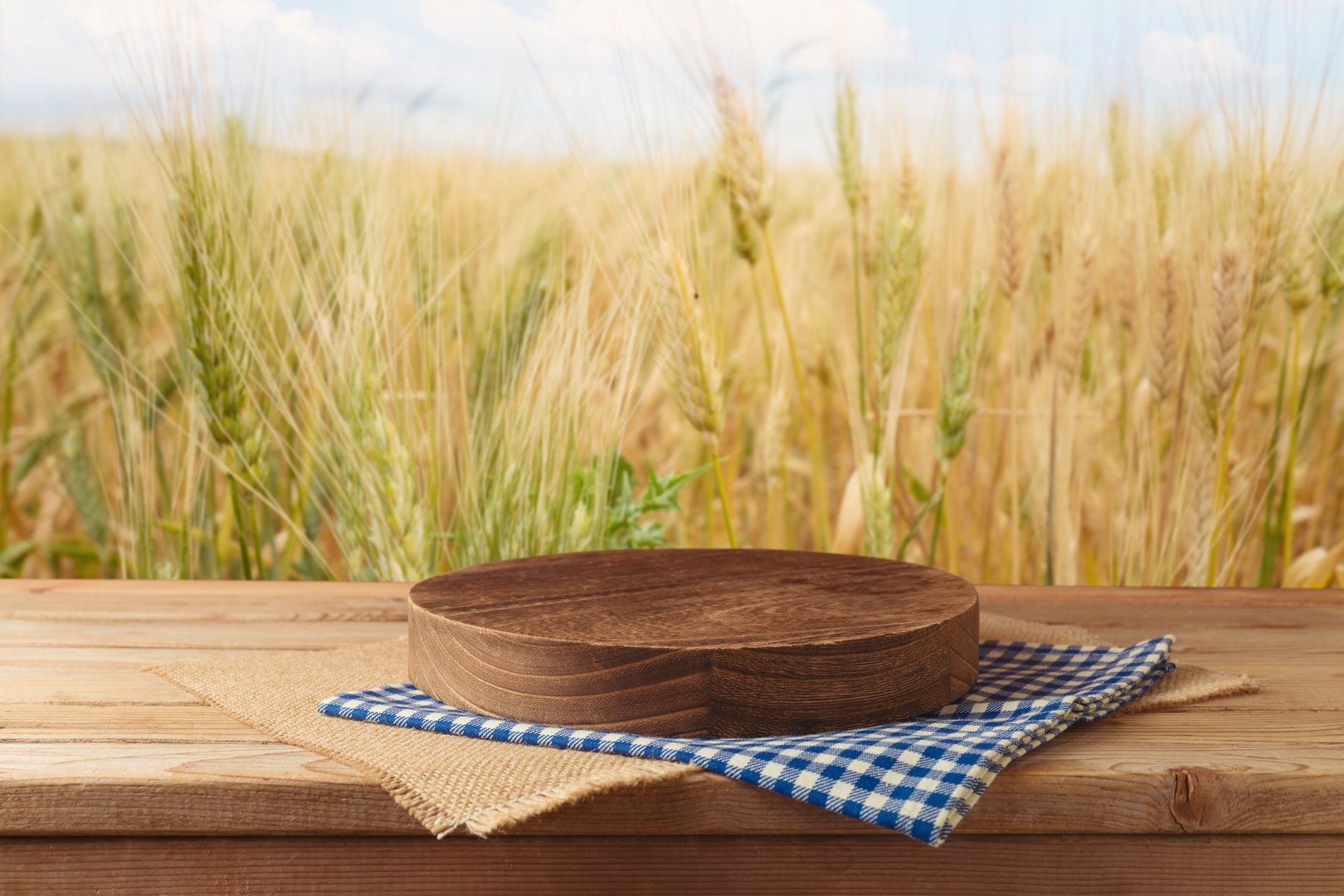 Empty wooden table with podium and tablecloth over wheat field blurred background. Shavuot holiday mock up for design and product display