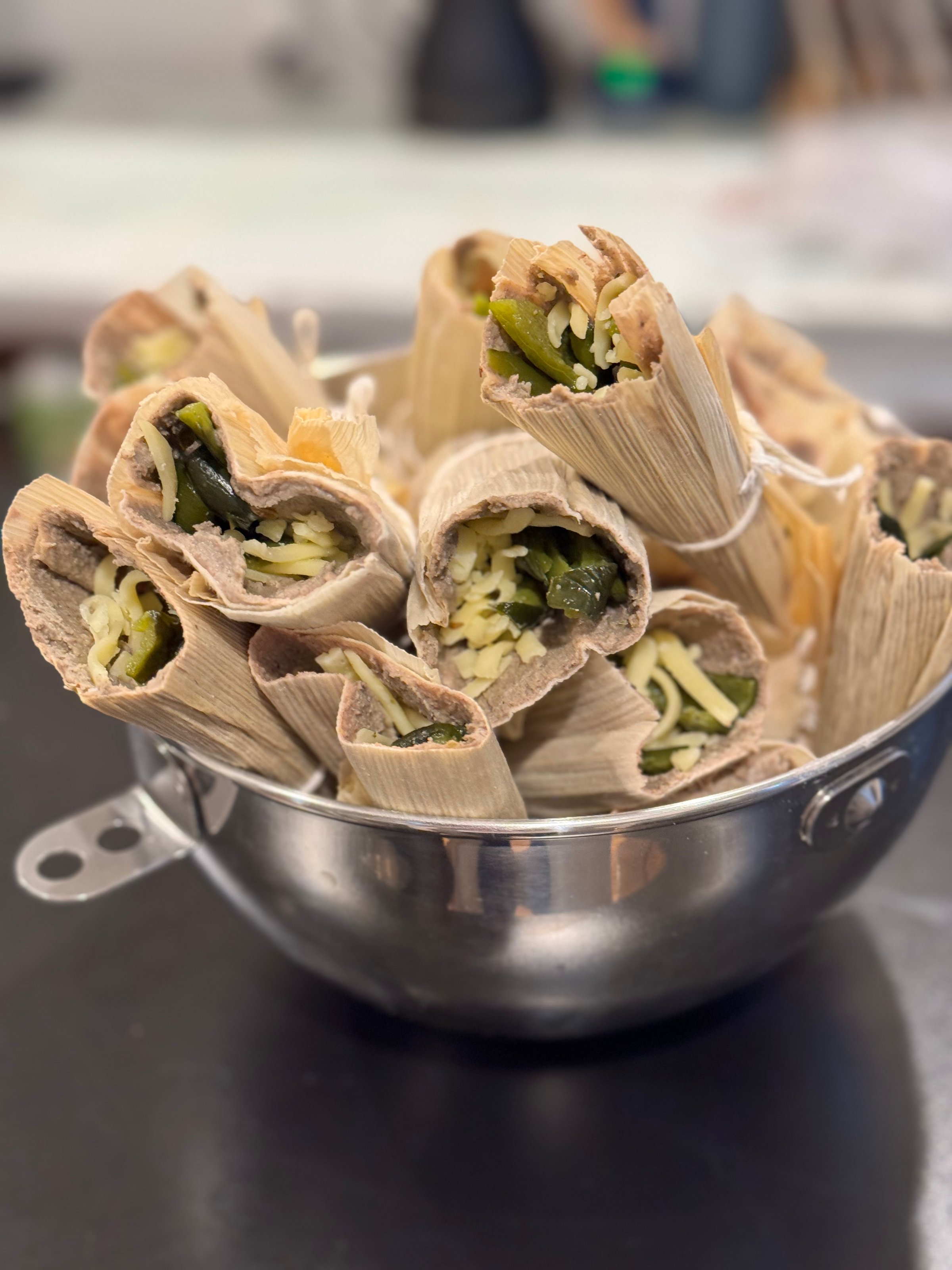 Tamales filled with cheese and green peppers, wrapped in corn husks, displayed in a metal colander.