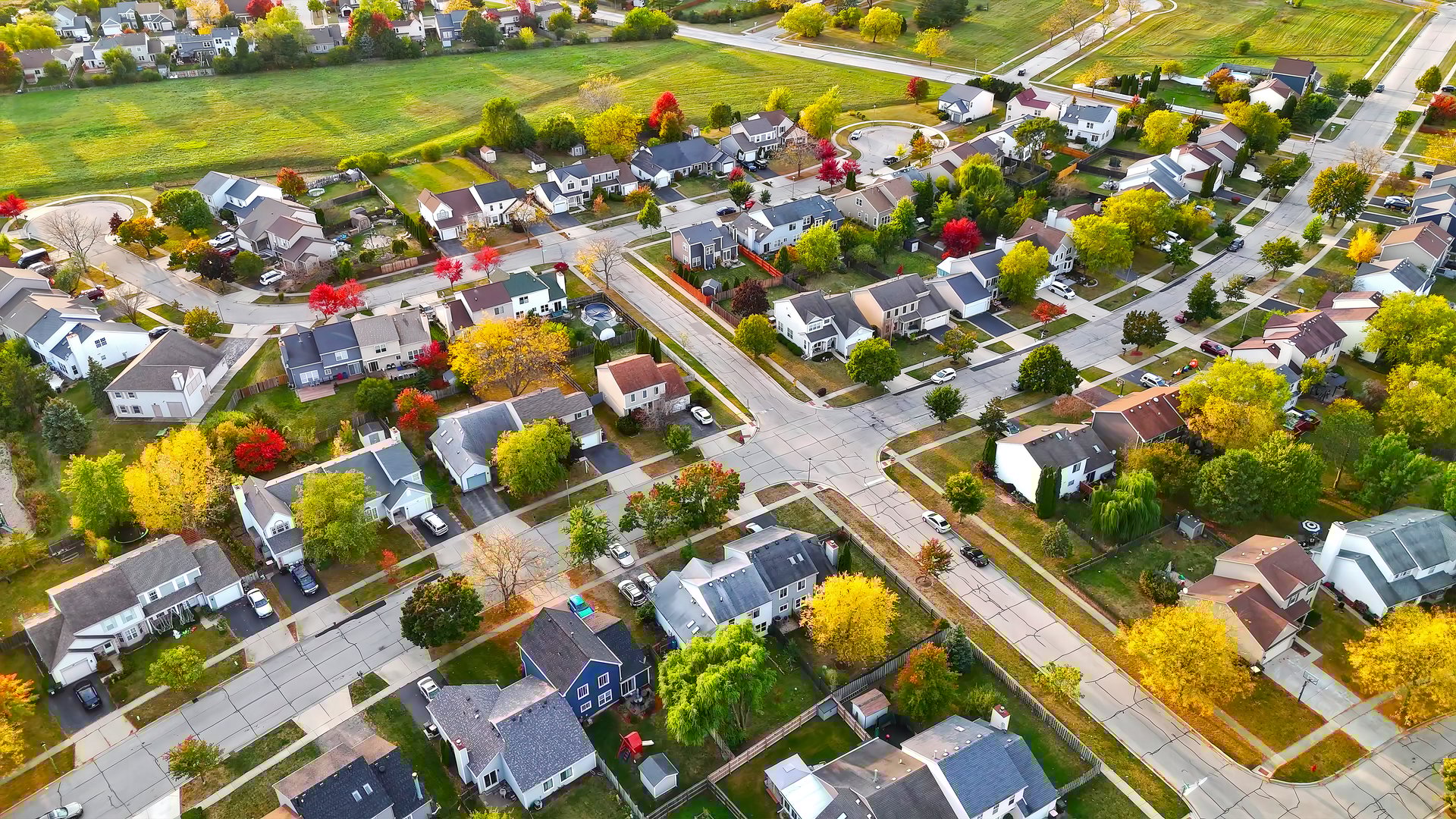 Aerial View of a Suburban Neighborhood in Autumn. A vibrant aerial view of a suburban neighborhood in autumn, showcasing colorful trees, neatly arranged houses, roads, and a nearby open green space under natural sunlight.