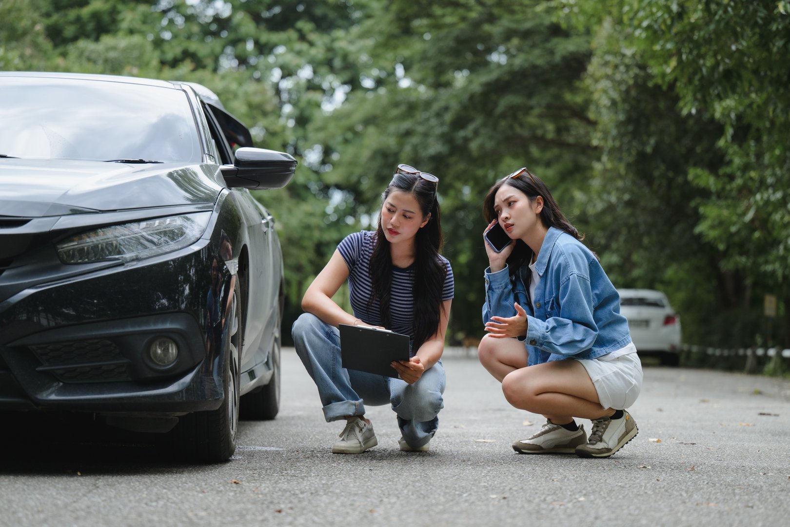 Two young women crouching near a damaged car, one holding a clipboard and the other making a phone call for assistance