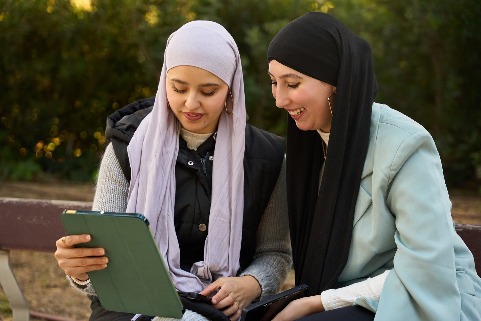 Two smiling young women wearing hijabs are sitting on a bench in a park, looking at a digital tablet together