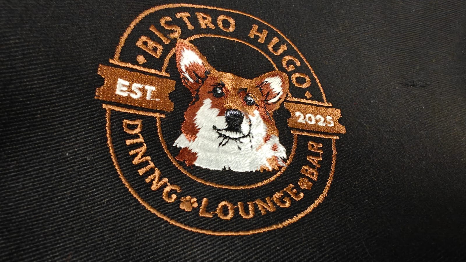 Embroidered logo featuring a corgi face with text: Bistro Hugo, Est 2025, Dining Lounge Bar, on a dark fabric background.
