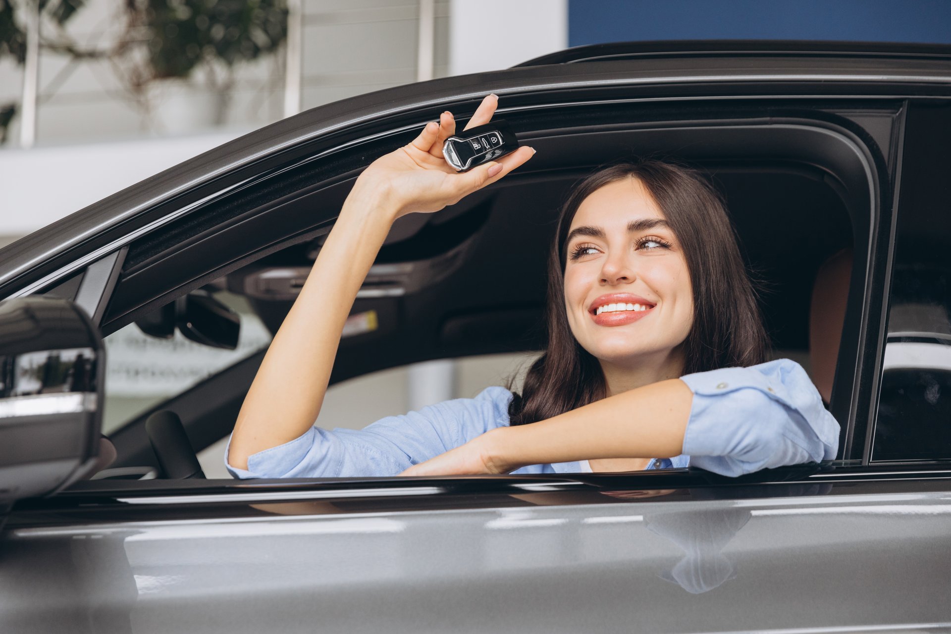 Happy smiling woman sitting in new car and holding keys in car dealership showroom