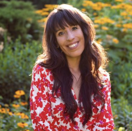 Woman with long dark hair wearing a red floral dress, smiling in a garden with yellow flowers.