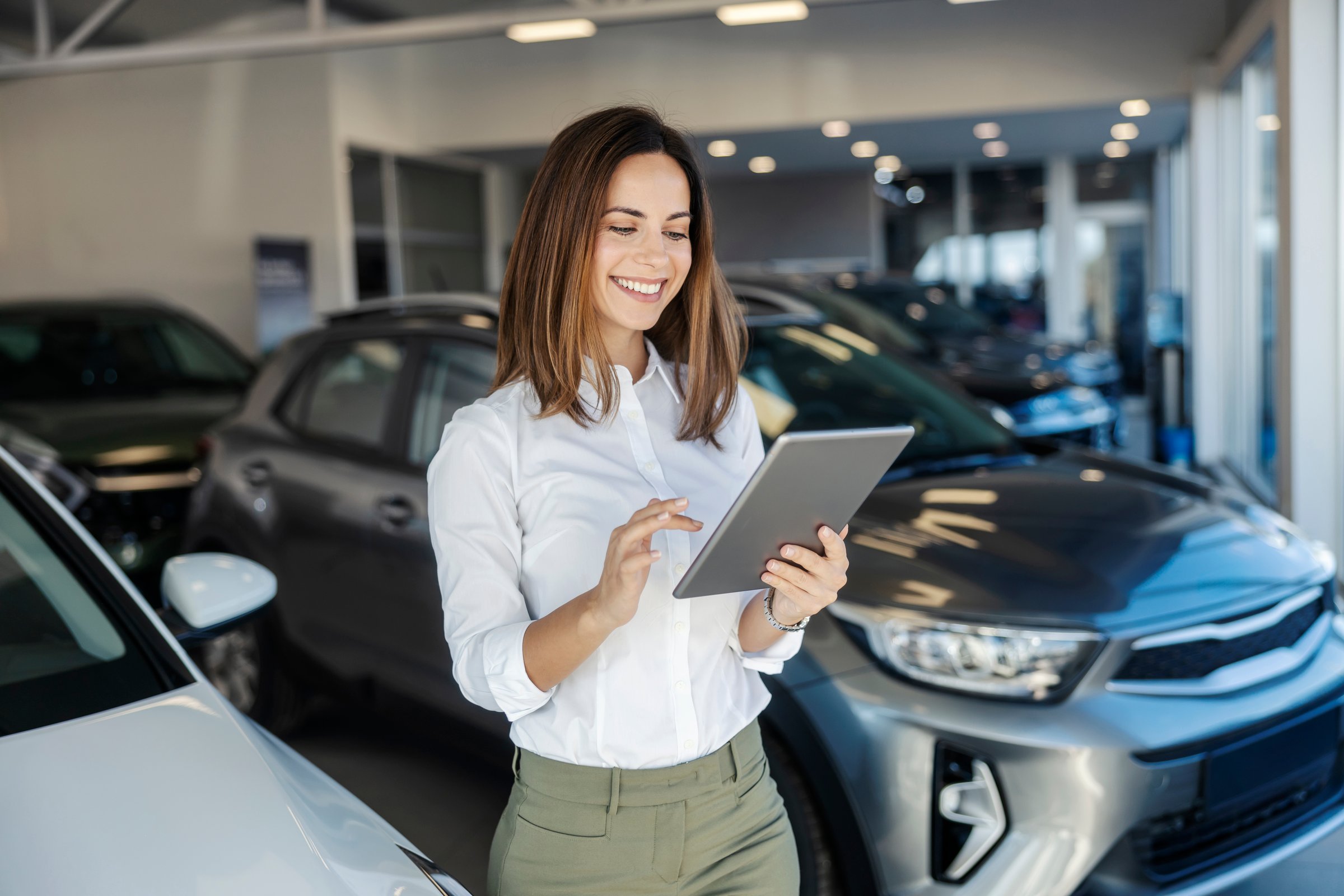 Portrait of happy professional female car dealer standing at car showroom with tablet in hands and scrolling on it.