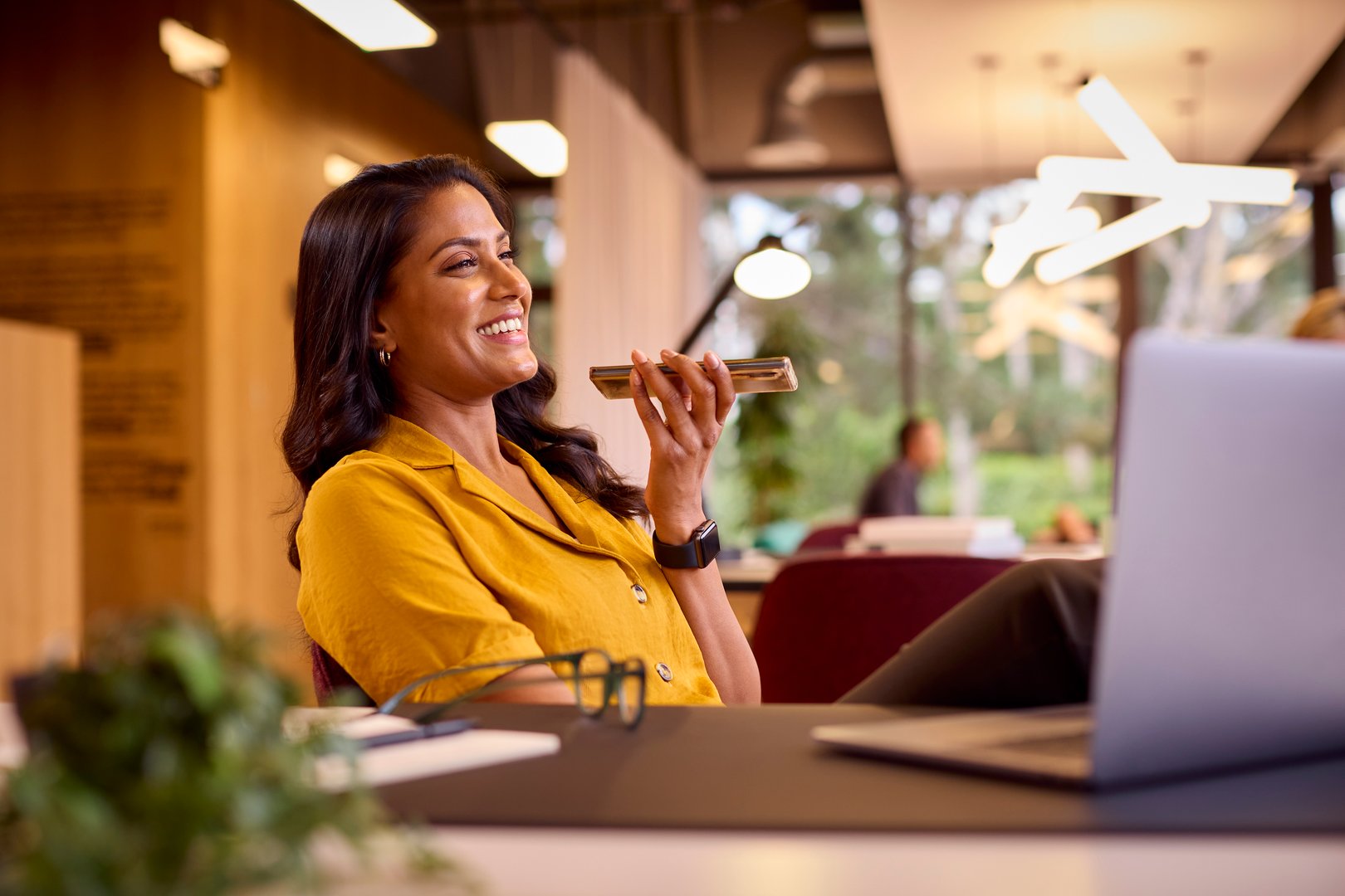 Mature Businesswoman Working On Laptop At Desk In Office Talking Into Mic Of Mobile Phone
