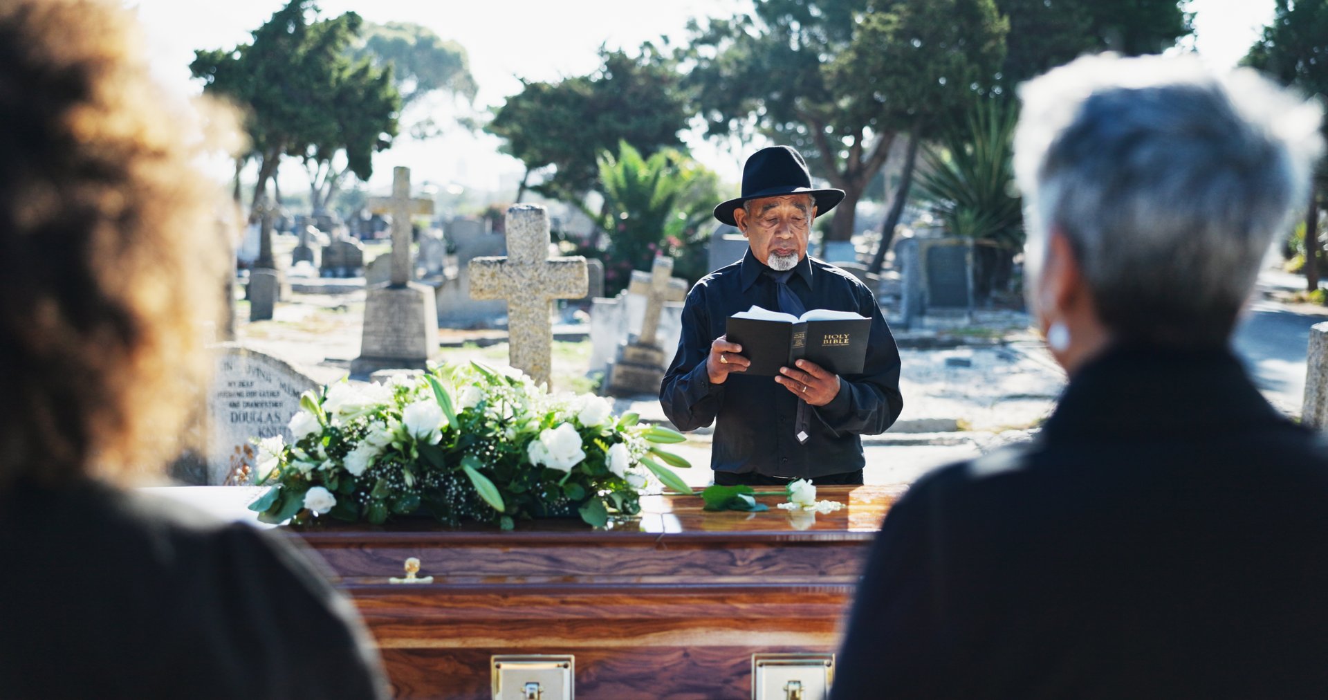 Bible, coffin and priest by graveyard with family for spiritual farewell, grief or loss. Religion, casket and pastor with people for memorial service at burial with holy book for blessing or goodbye.