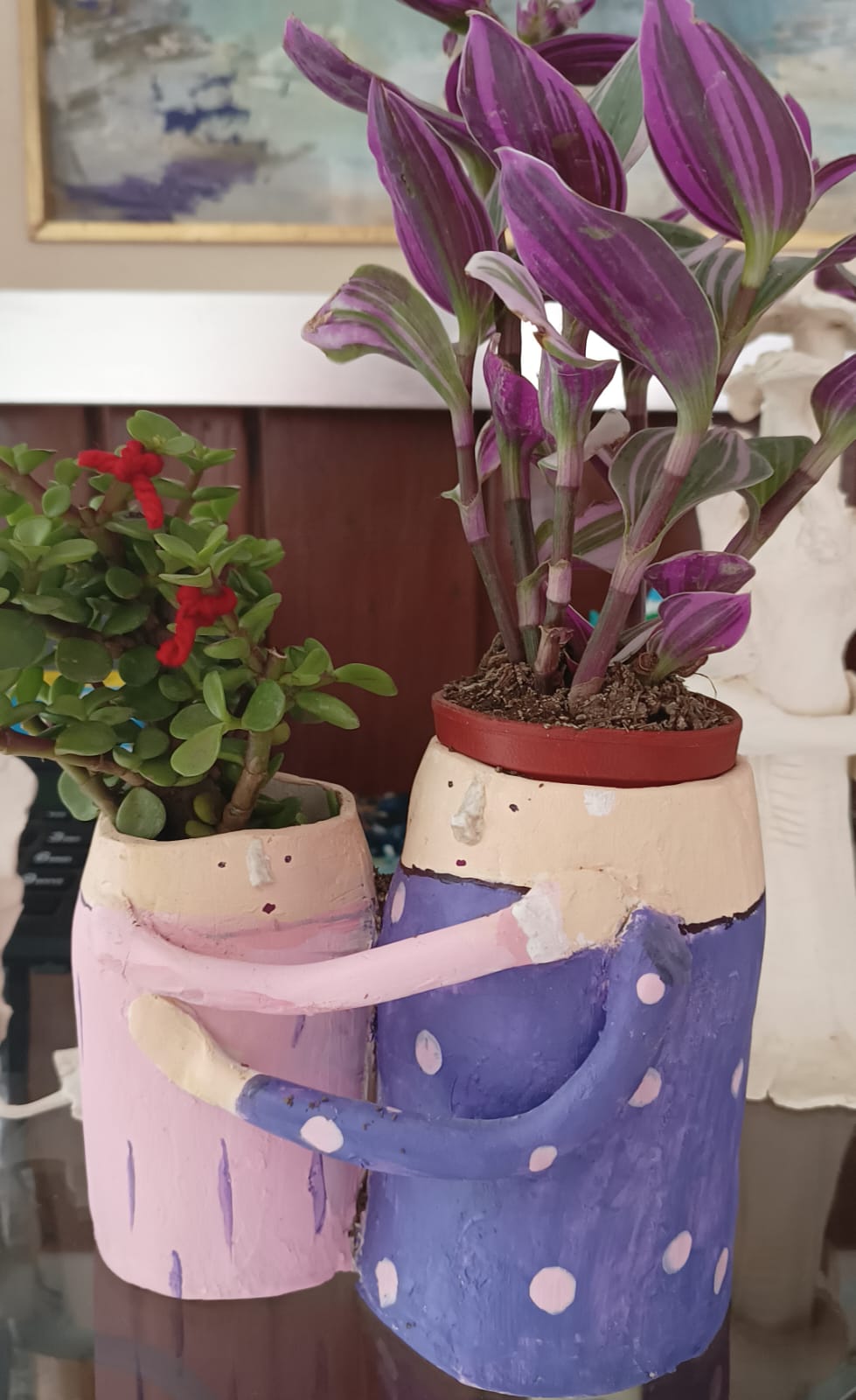 Two colorful plant pots with face designs, one with leafy green plants and the other with purple foliage, on a table.