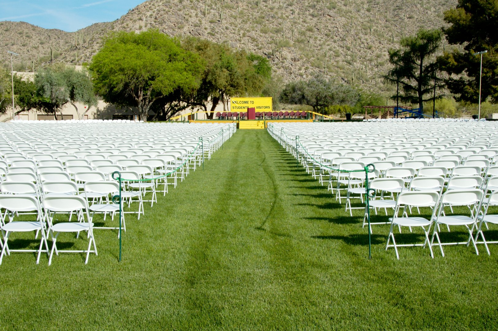 Rows of white folding chairs on a grassy field facing a stage with a yellow sign and mountains in the background.