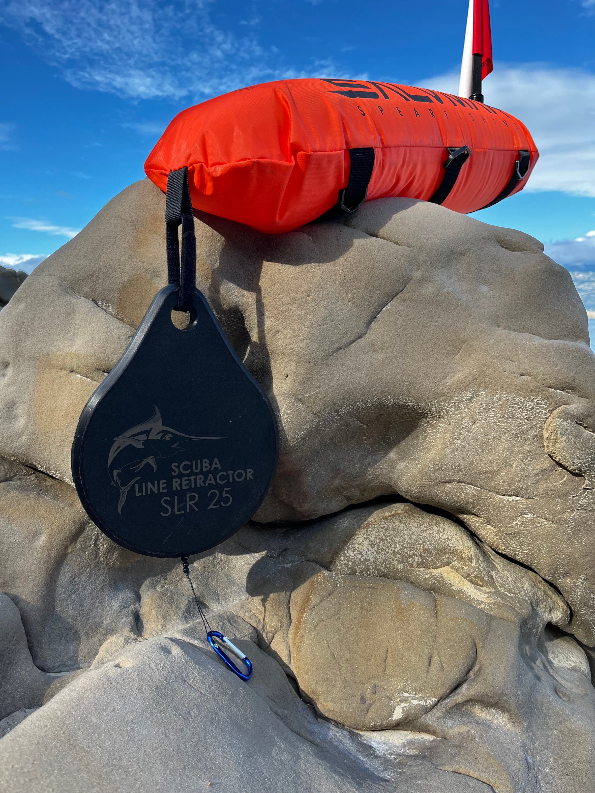 A scuba line retractor and orange float resting on a rocky surface against a blue sky.