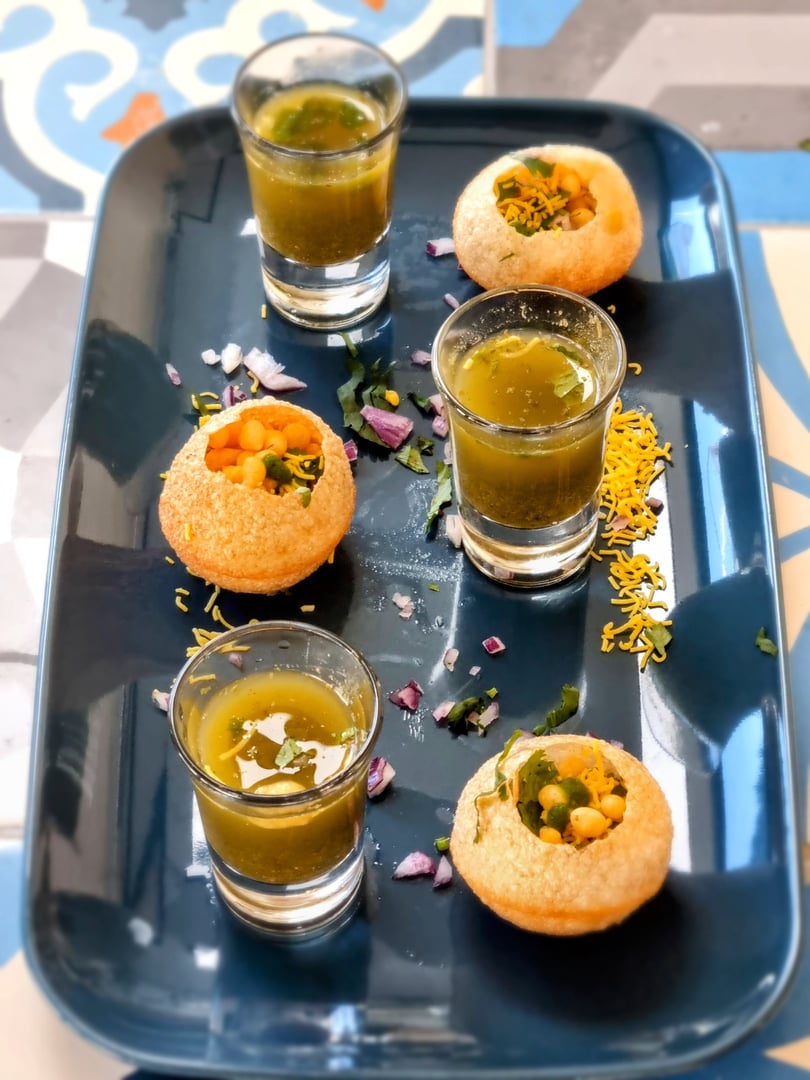 Pani puri with spicy water in shot glasses on a dark tray, garnished with sev, herbs, and chopped onions.