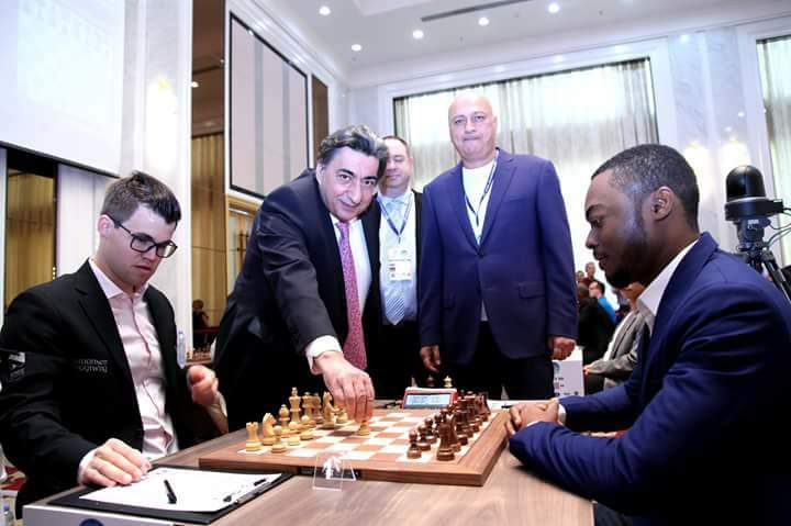 A chess player in a suit makes the opening move while others, also in suits, watch. Two seated players are ready to start the game.