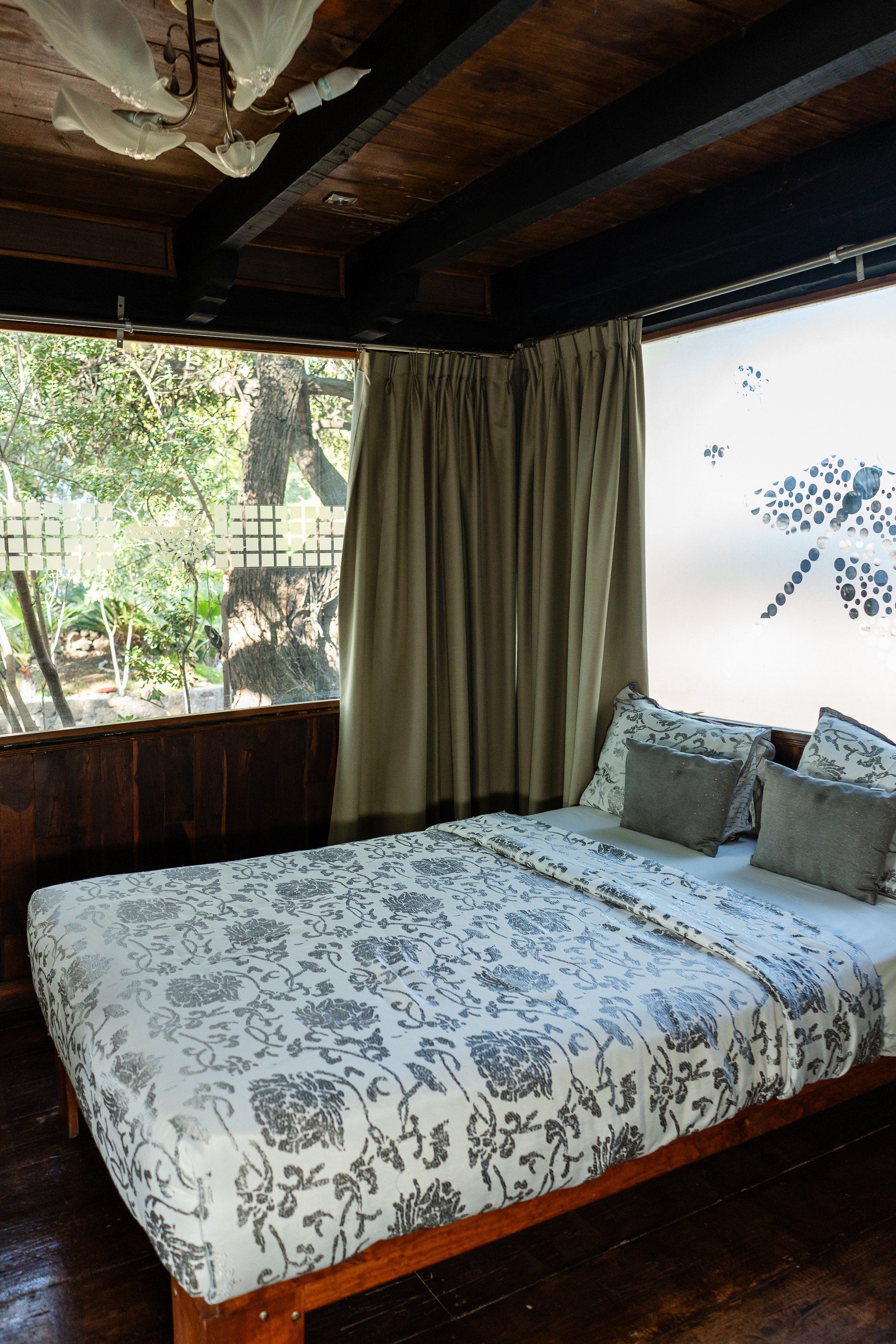 Cozy bedroom with a floral-patterned bedspread, wooden decor, large window with curtains, and a frosted glass panel.