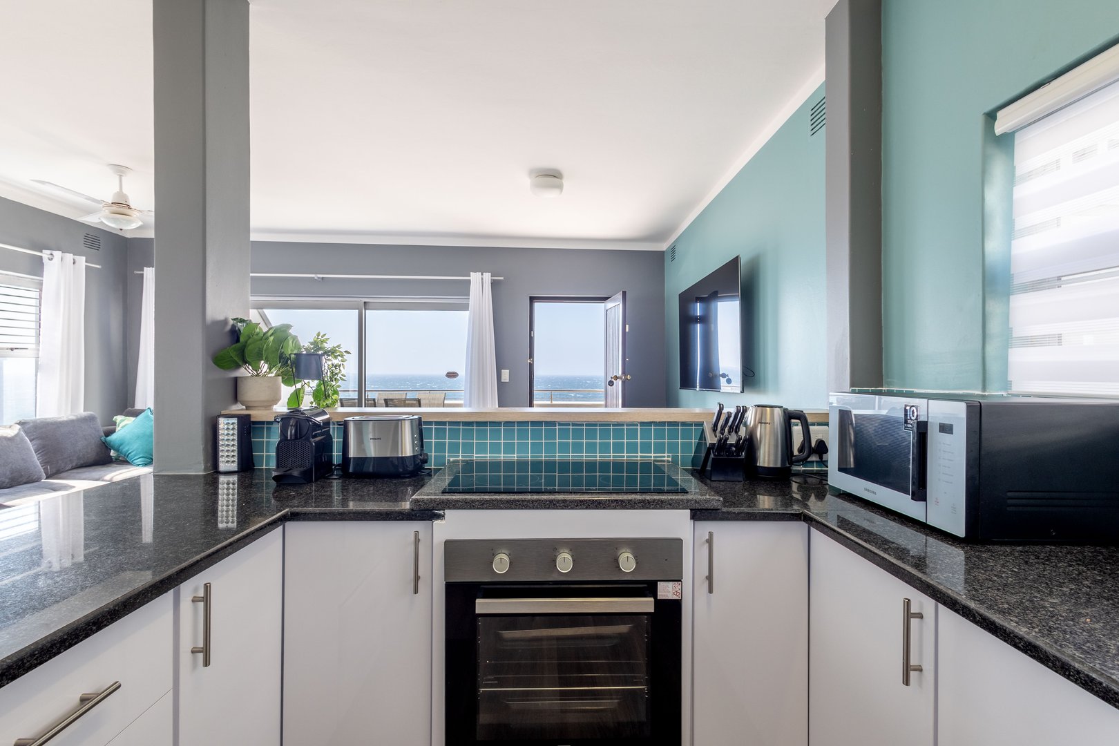 Modern kitchen with black granite countertops, oven, toaster, microwave, and ocean view through large windows.