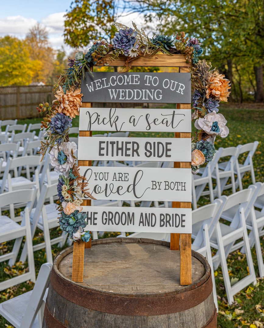 A wedding welcome sign on a barrel with floral decorations and white chairs in an outdoor setting in Elkhart County, Indiana