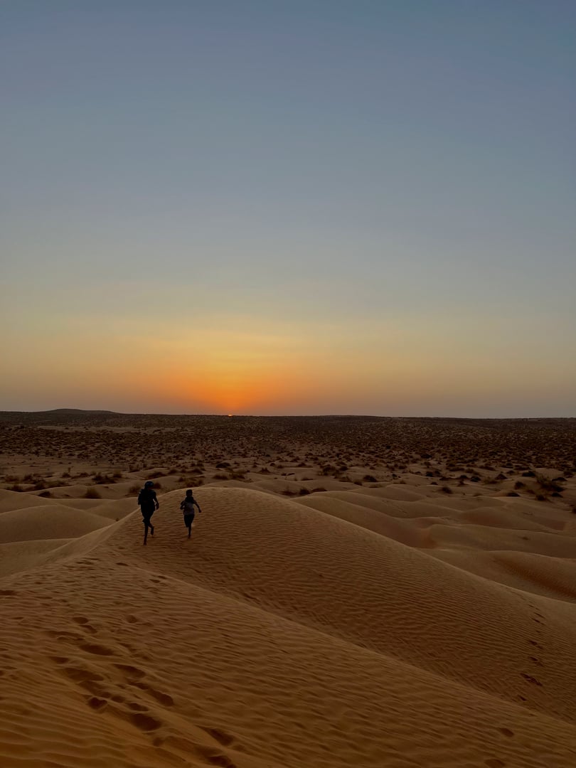 Two children playfully run across the golden sand dunes of Douz, Tunisia, enjoying the tranquil evening as the sun sets in the background, creating a serene atmosphere.