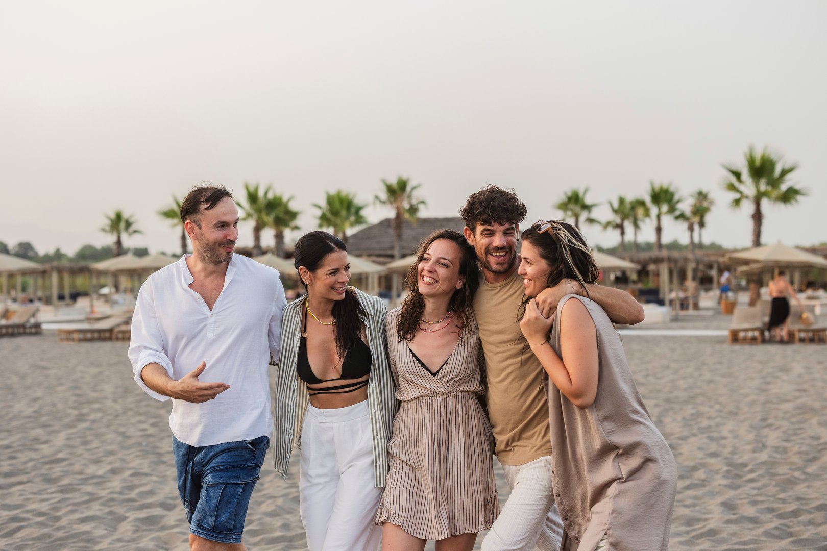 Five cheerful young adults walking along the beach at sunset, savoring their vacation while sharing joyful moments filled with laughter and friendship
