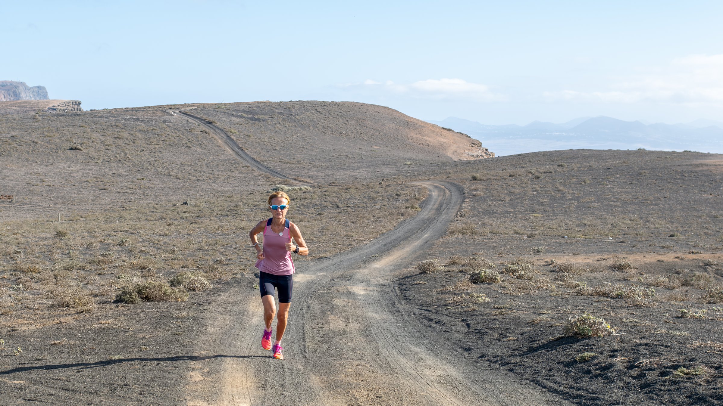 A woman in athletic wear running on a dirt track in a barren desert landscape under a clear sky.