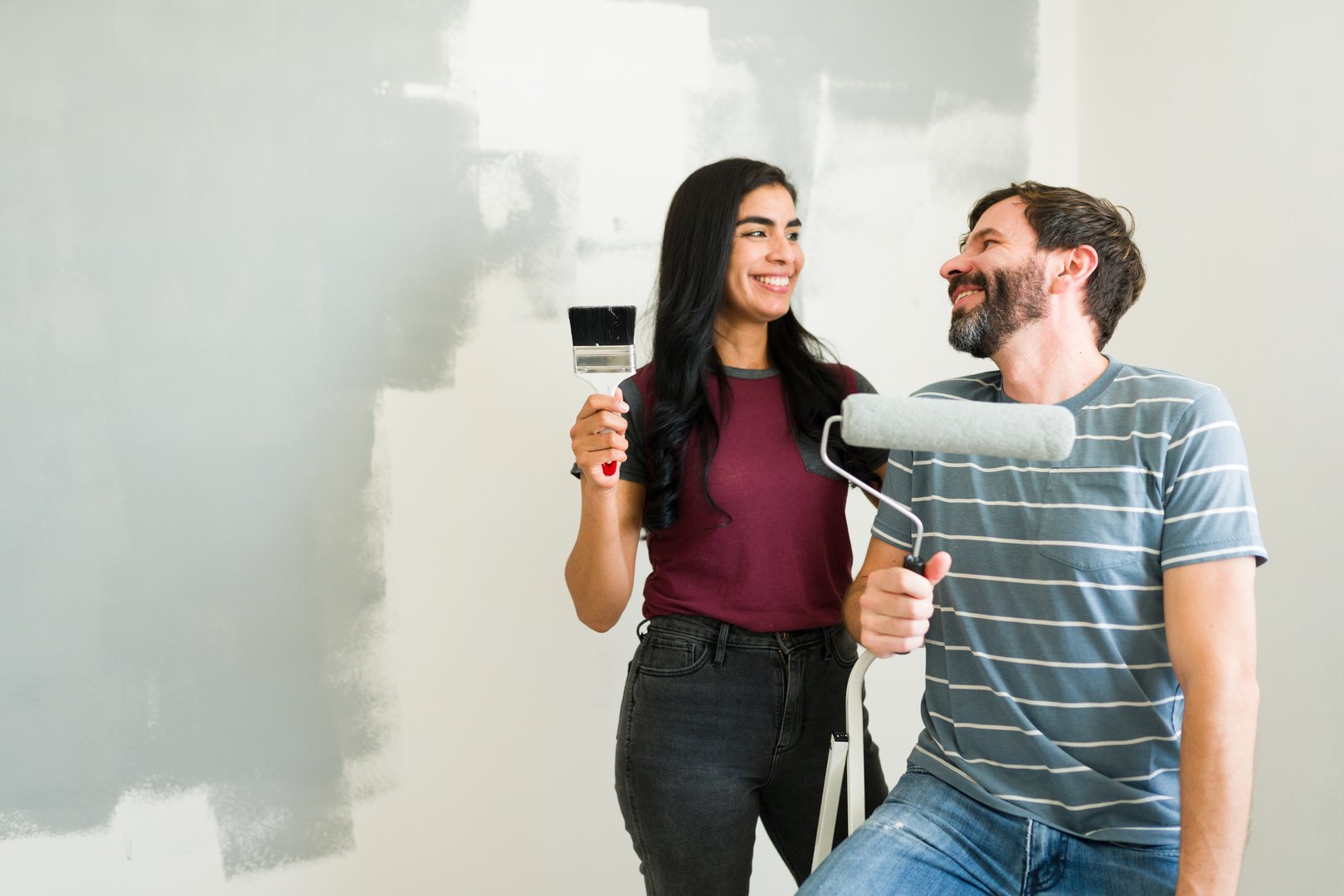 Young couple smiling and looking at each other while holding painting tools, taking a break from painting the walls in their new home