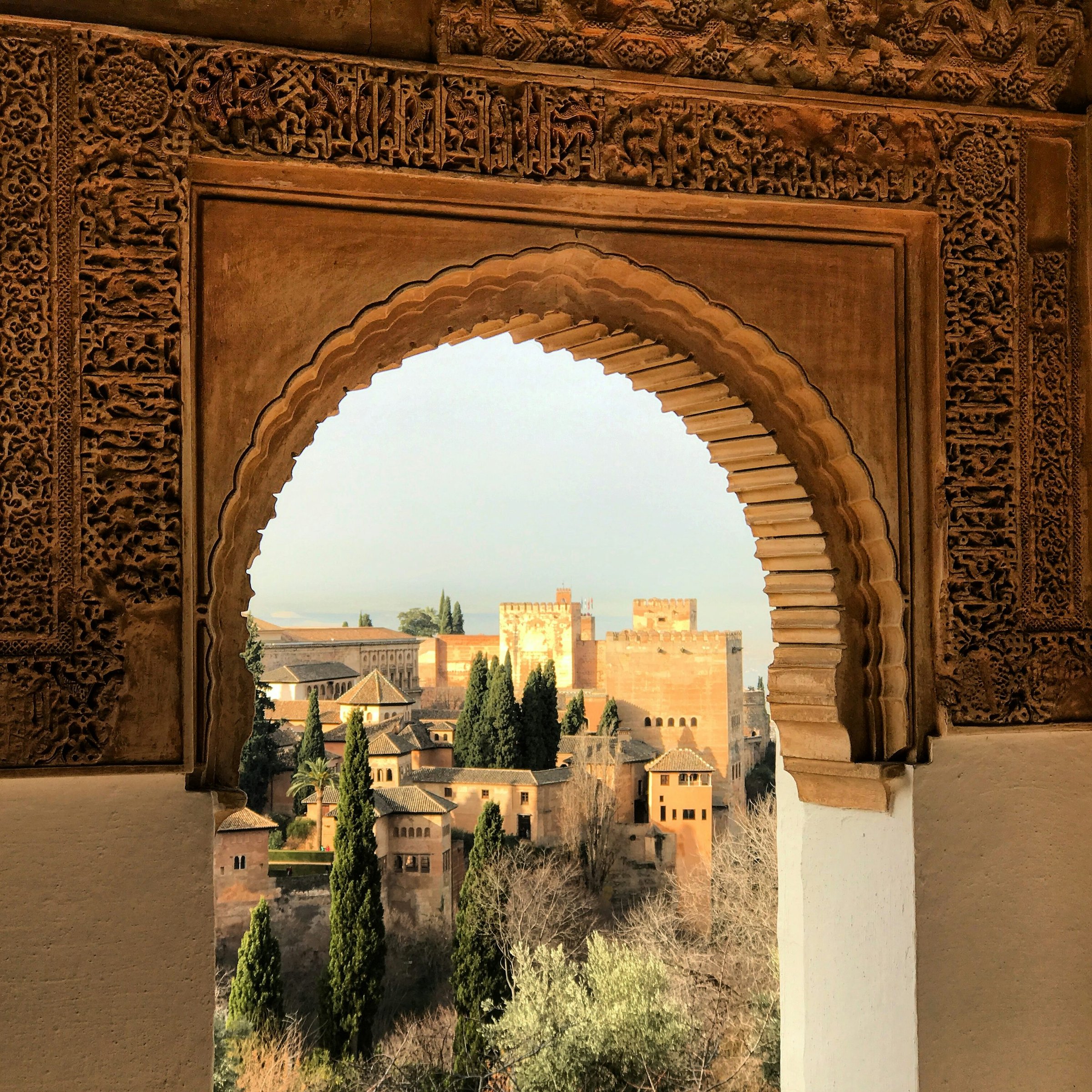 View of the Alhambra through an ornate arch with intricate carvings, surrounded by lush greenery and a clear sky.