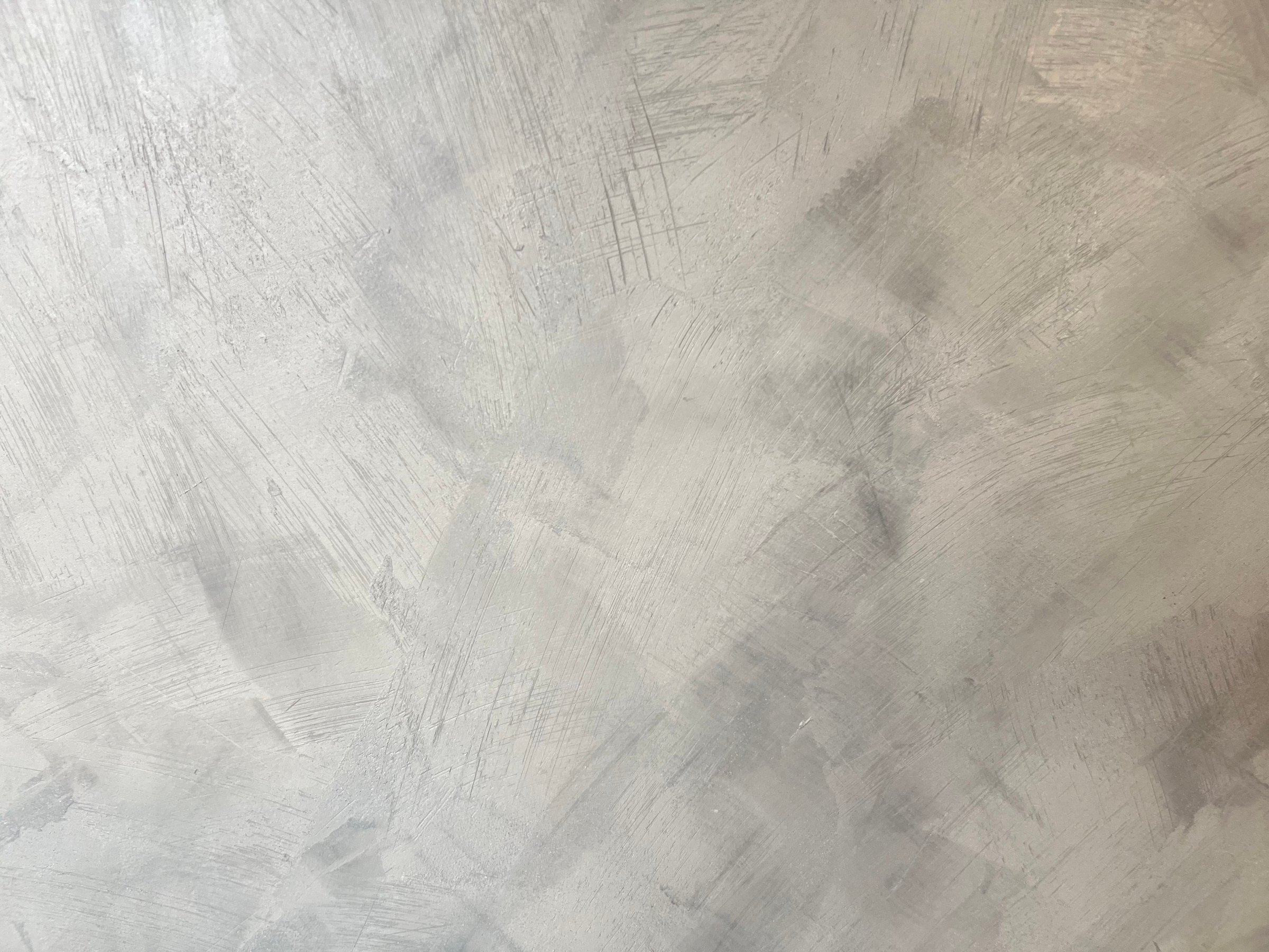 A close-up of a textured concrete wall with a rough, abstract pattern. The surface features light gray tones with visible brush strokes, scratches, and irregular patterns, creating a modern and industrial aesthetic. This texture is commonly used in interior design, architecture, and backgrounds for creative projects.
