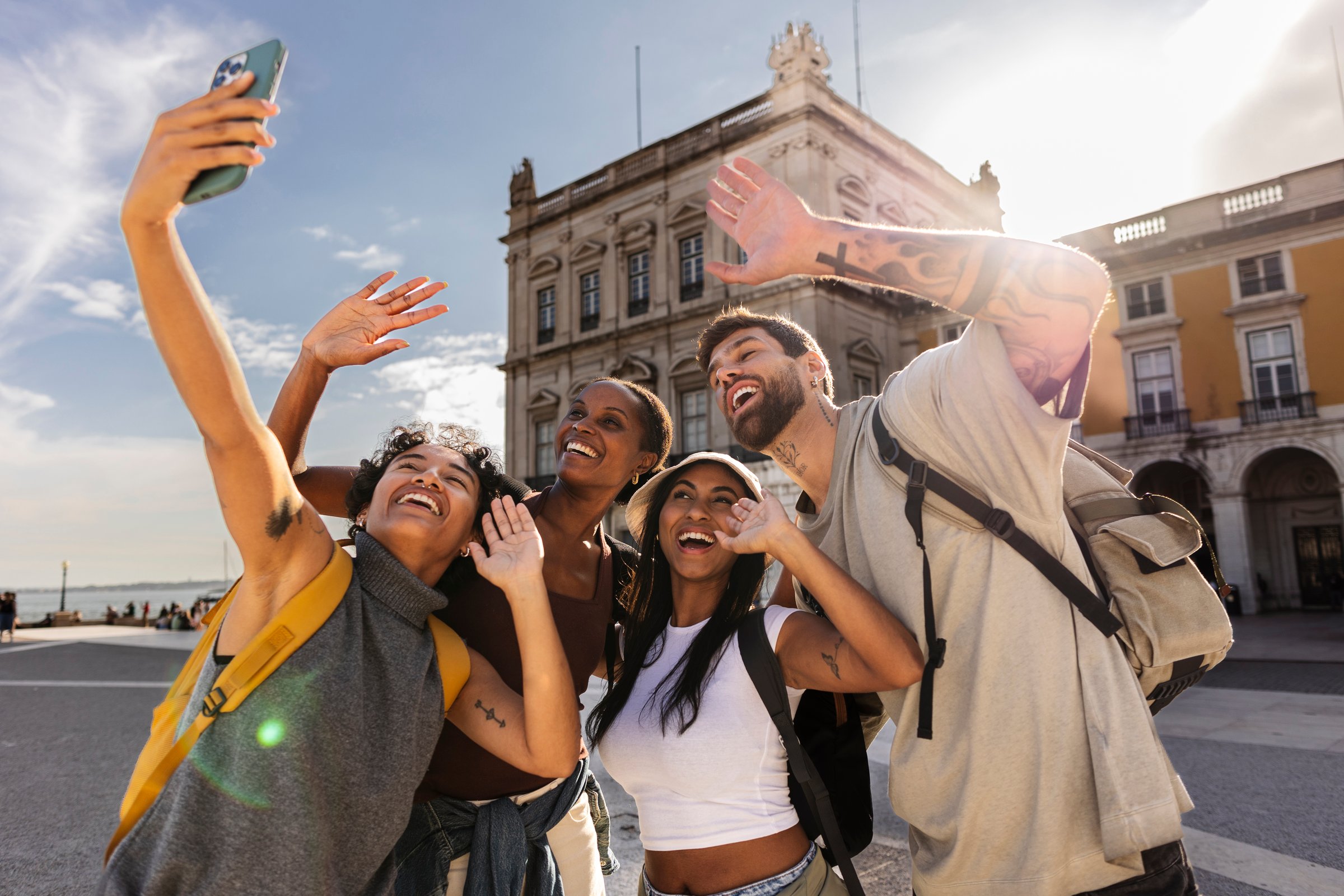 Four cheerful young tourists, two men and two women, are taking a selfie with a smartphone in lisbon, portugal, waving their hands and smiling happily