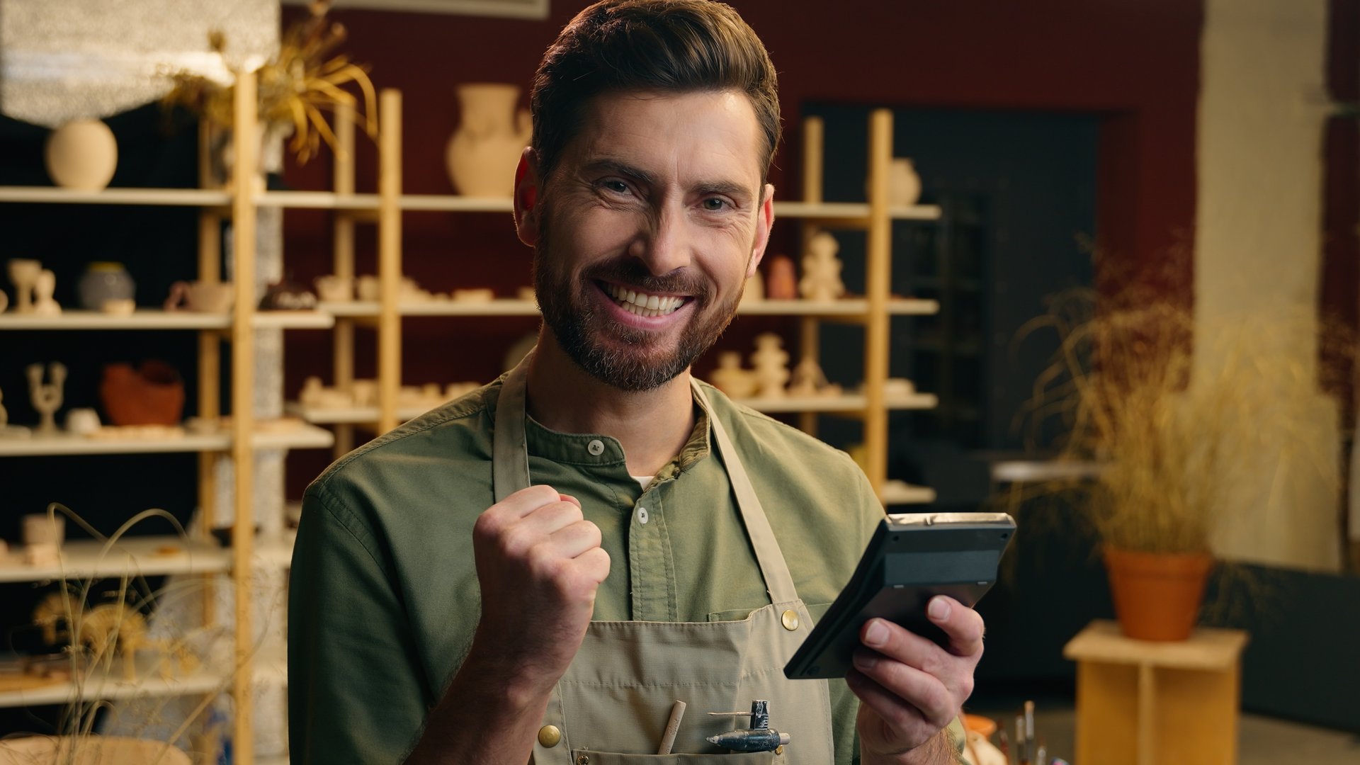 Happy successful Caucasian small business owner man male guy businessman salesman potter holding calculator calculating finance budget success growth money clench fist gesture win luck indoor workshop. High quality 4k footage