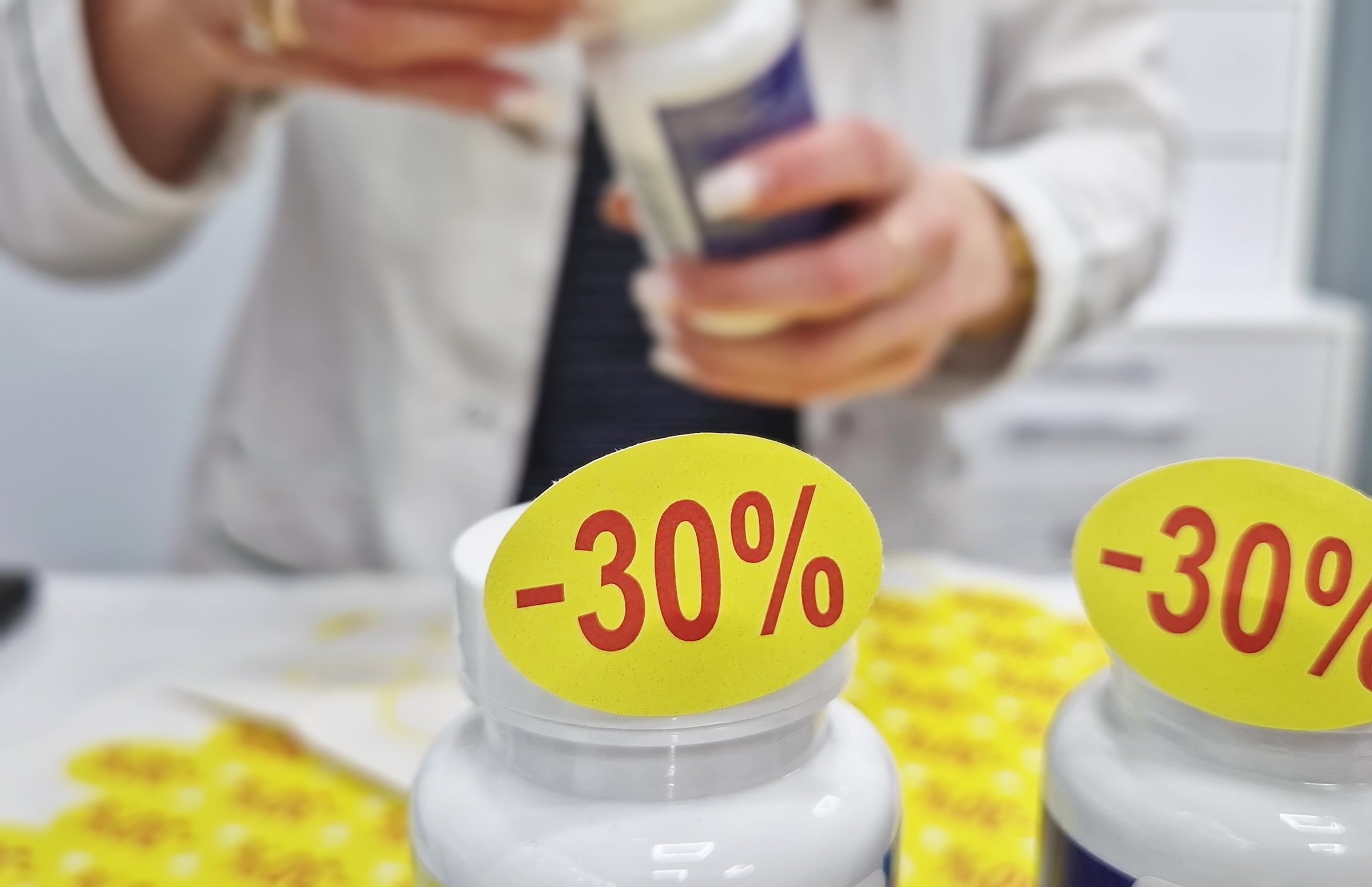 Young woman pharmacist wearing white uniform attaching discount stickers to medicine items from stocked pharmacy or medical supply store. Marketing advertising with 30% cutprice sale at drugstore