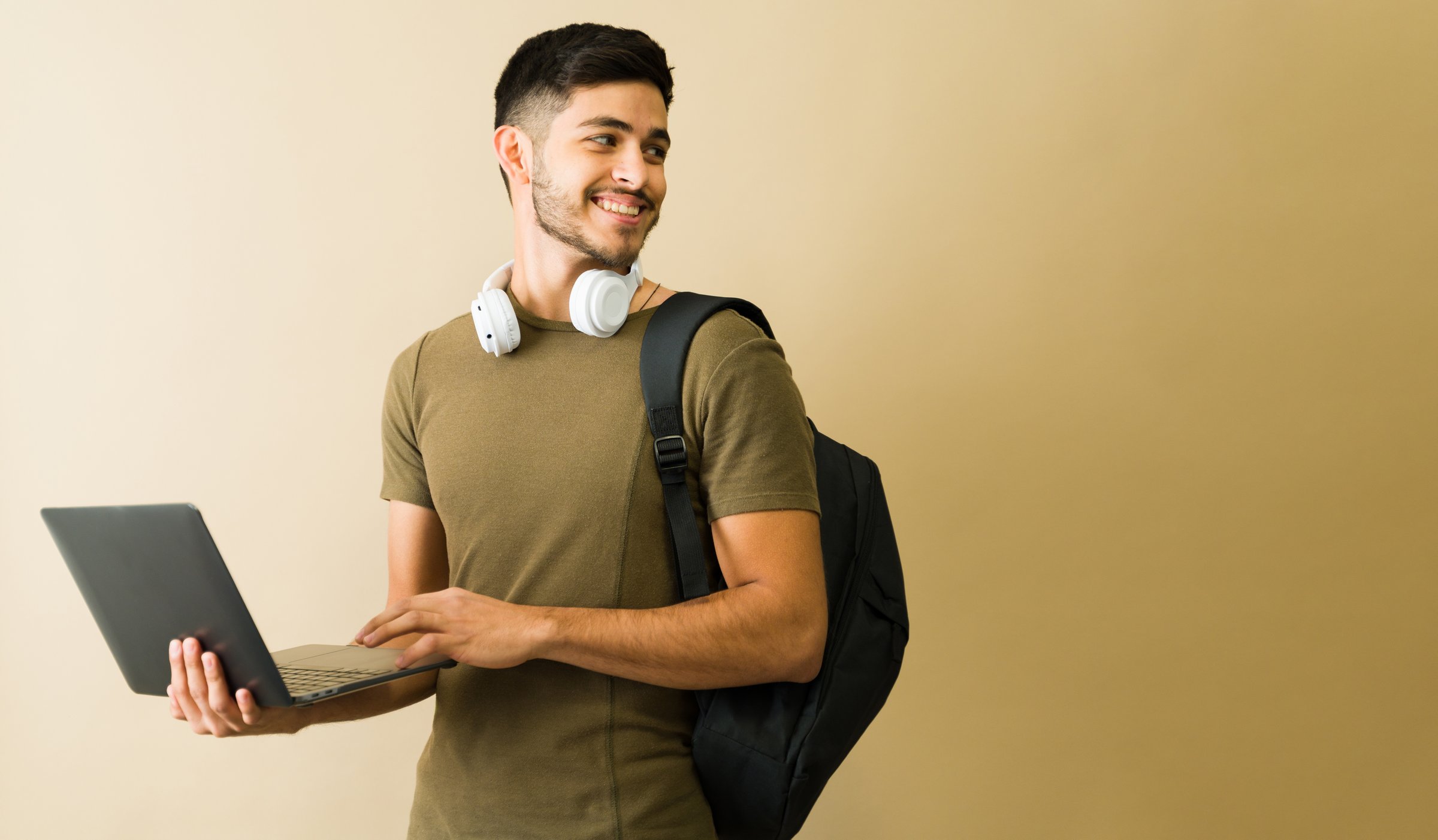 Happy young man wearing headphones and carrying backpack using laptop while walking and looking away