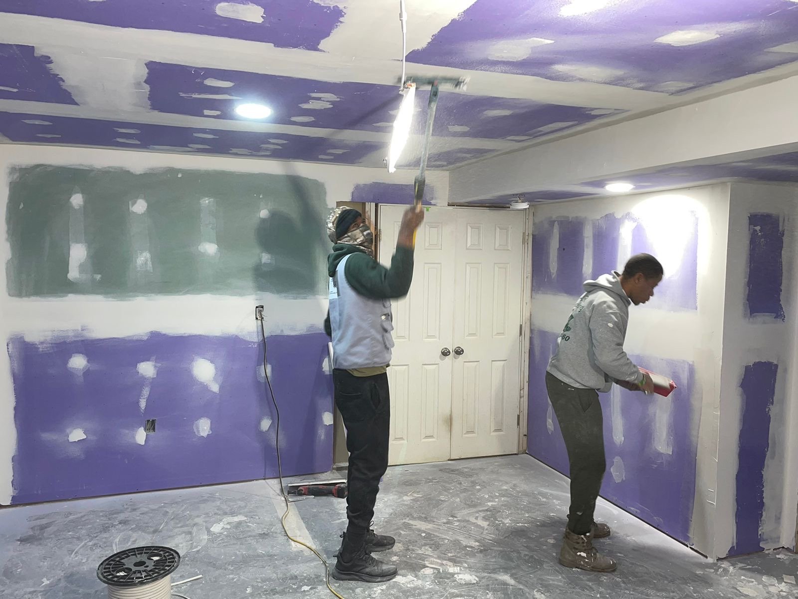 Two people painting walls and ceiling in a room with drywall patches and purple paint, working under bright lighting.