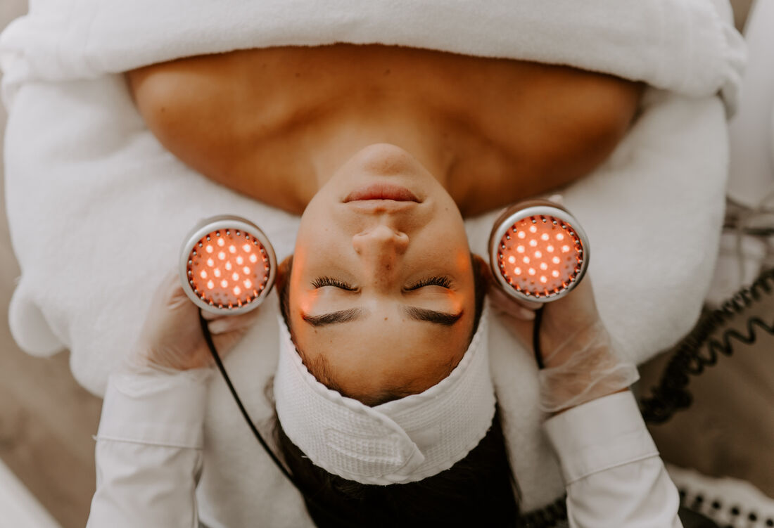 A woman receiving a facial treatment with red light therapy devices held near her face by a technician.