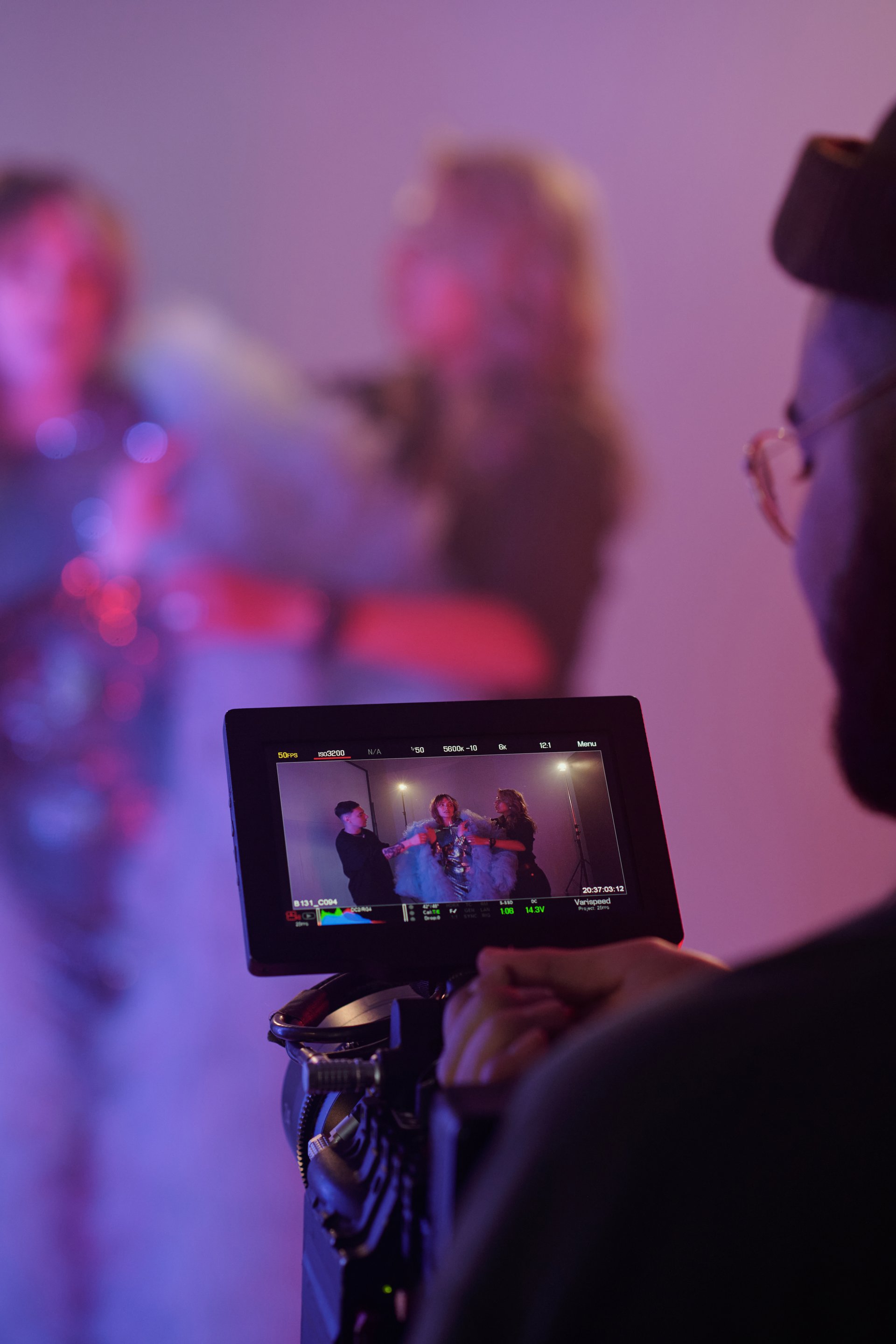 Vertical closeup of director watching camera monitor while filming styling scene with actress and assistants in studio under purple lighting