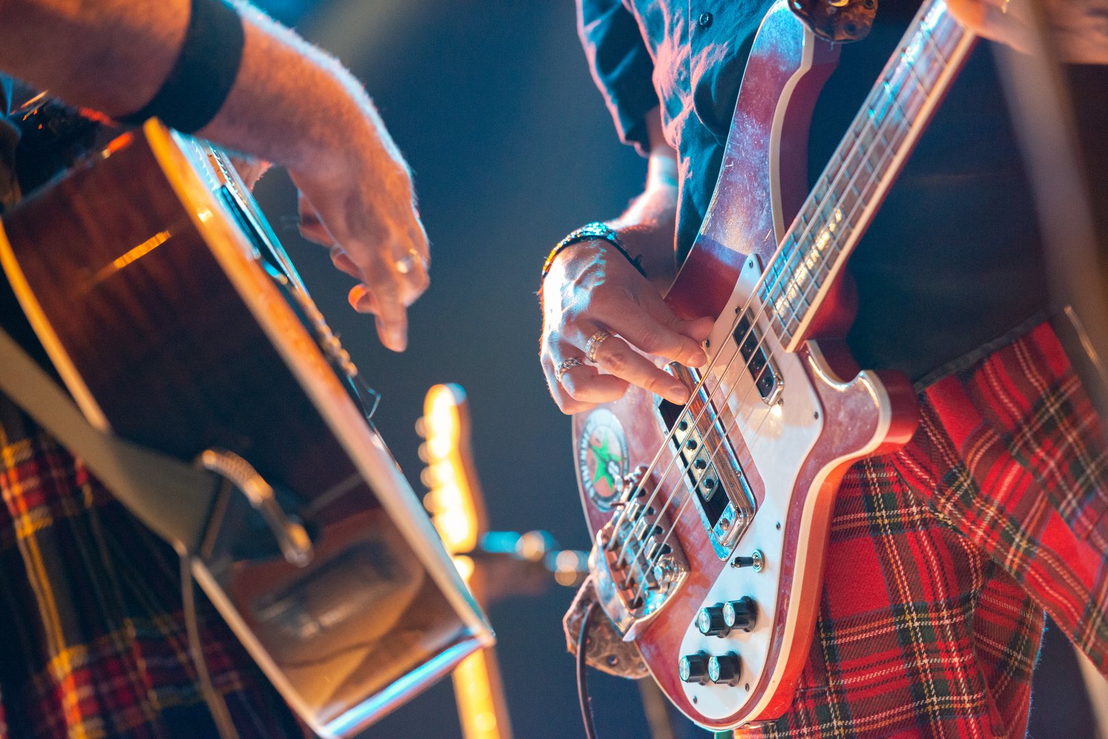 Close-up of musicians playing electric bass and acoustic guitar, wearing red plaid clothing, with stage lights in background.