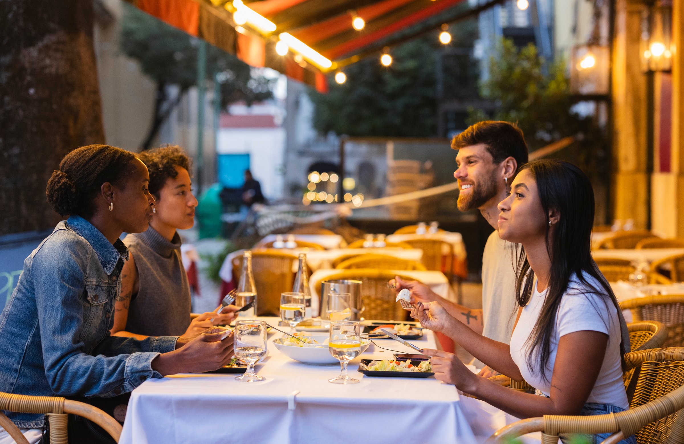 Four friends sitting around a table outside a restaurant in lisbon, portugal, enjoying a delightful meal and engaging in lively conversation under the evening lights of the vibrant city