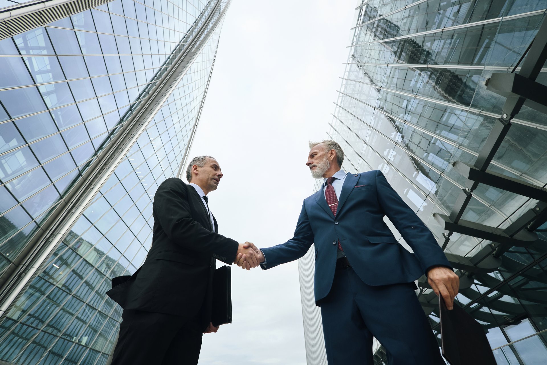 Upward view of two middle aged Caucasian men shaking hands outdoors between modern glass skyscrapers, both wearing business attire, engaging in professional agreement or partnership
