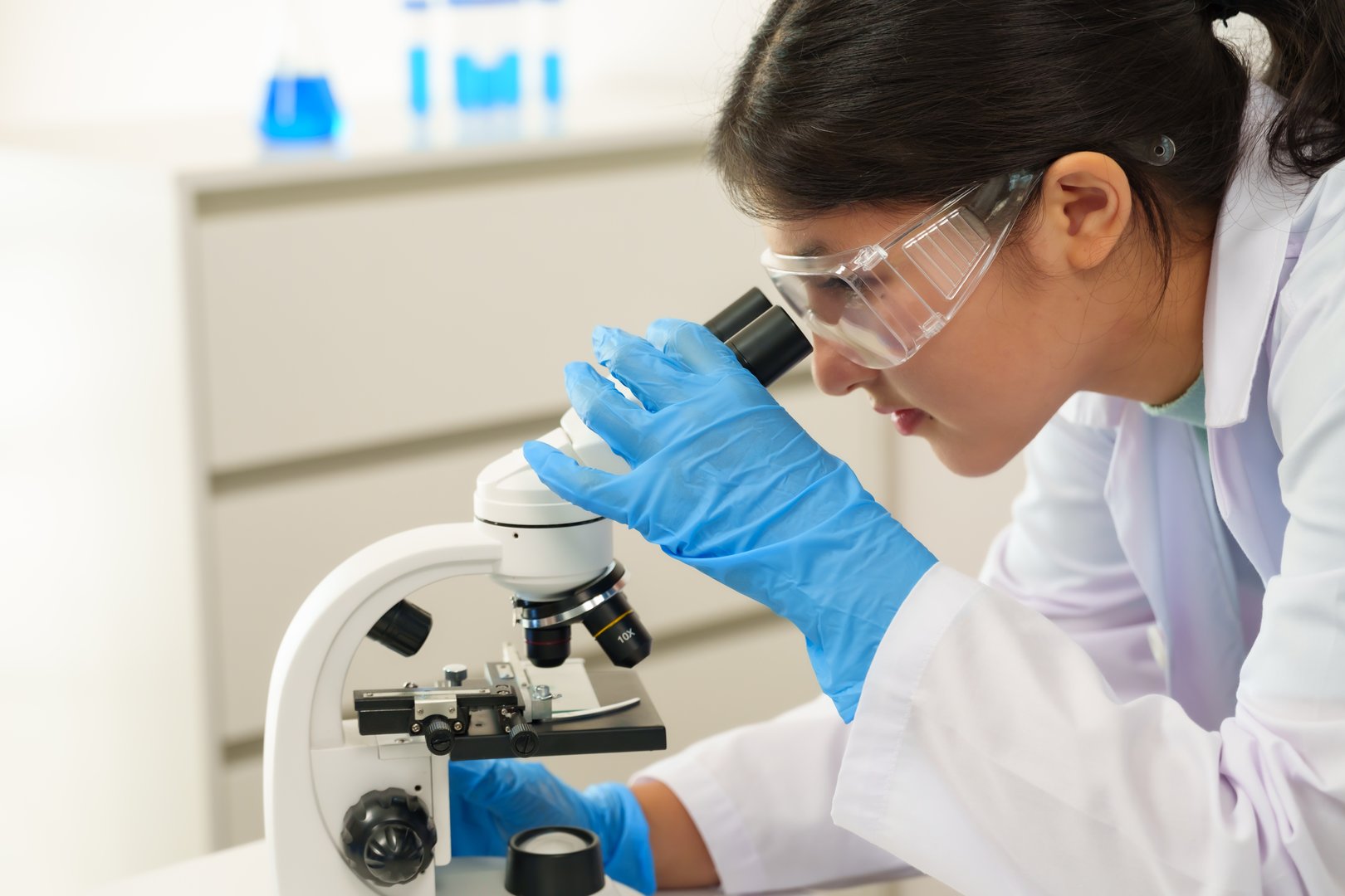 Asian female student wearing lab coat and safety goggles closely examines specimen under microscope in science classroom, demonstrating focused attention during exciting laboratory experiment.