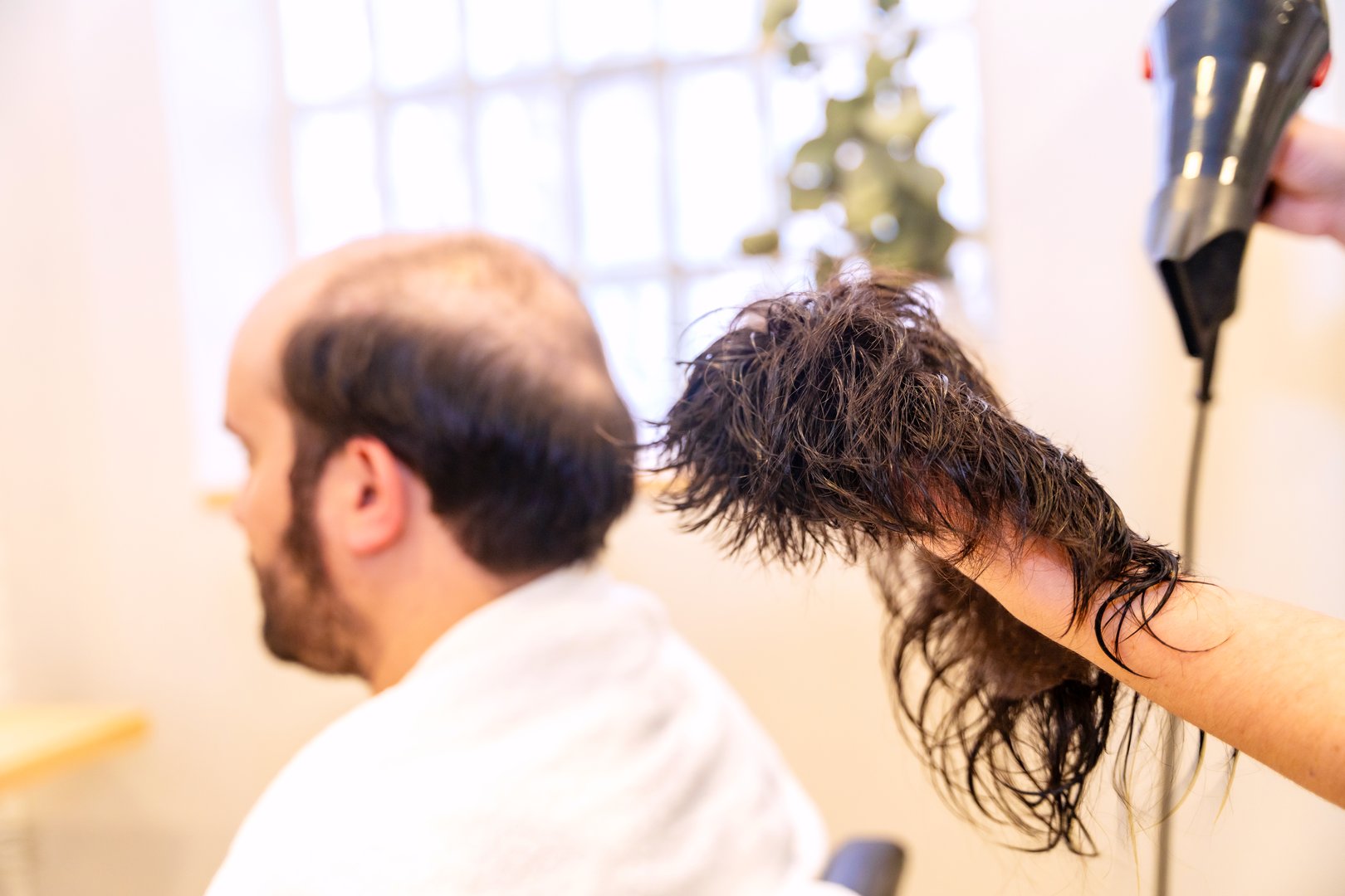 Bald man waiting while hairdresser cleaning and drying his capillary prosthesis in the salon